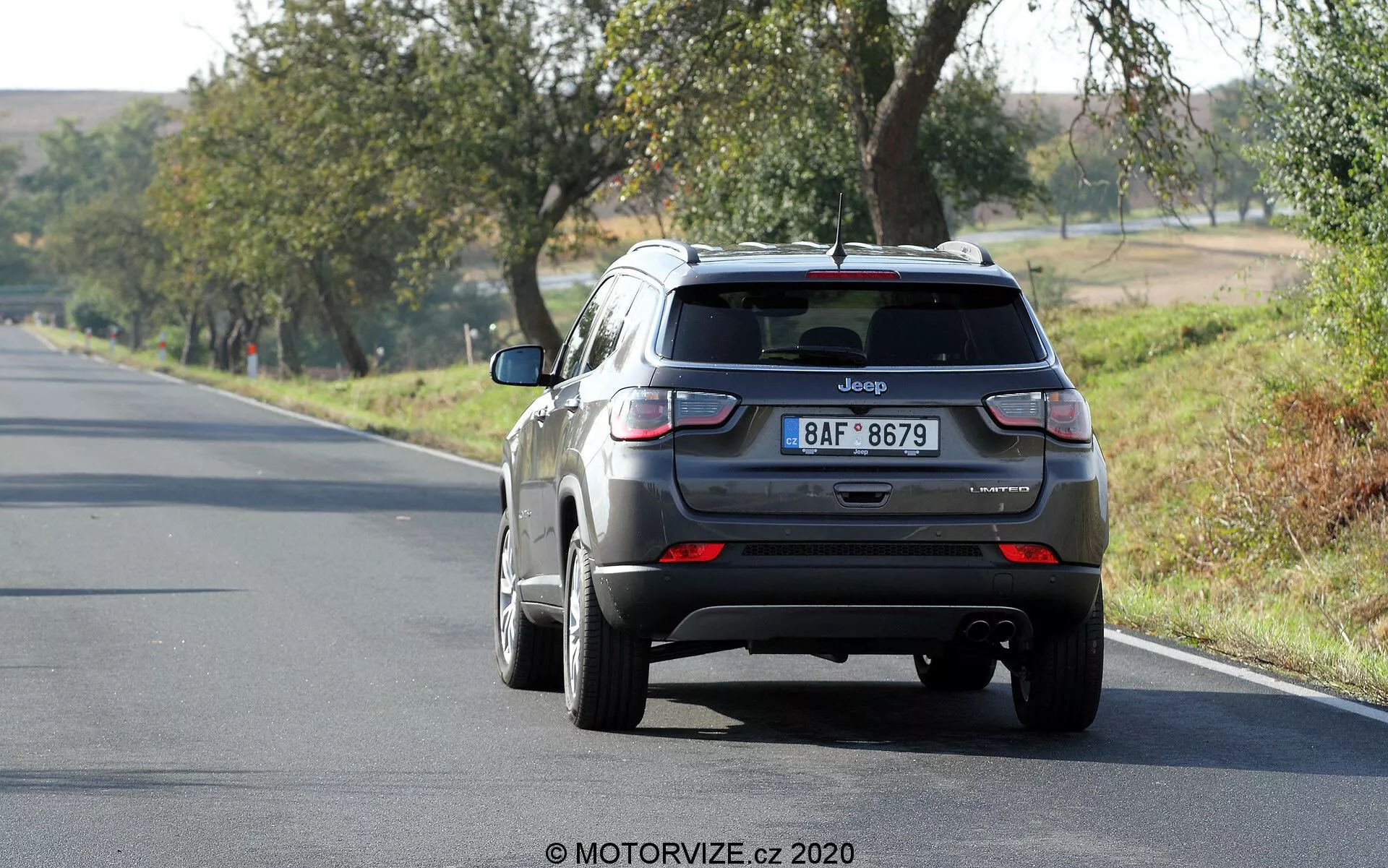 Vue arrière d'un SUV Jeep Compass 2017 gris métallique roulant sur une route à deux voies, légèrement à droite du cadre. Le véhicule, sur la voie de droite, présente des caractéristiques visibles telles qu'un hayon avec un badge Jeep et une plaque d'immatriculation, une lunette arrière avec un petit becquet, des feux arrière avec des sections rouges et blanches, un pare-chocs arrière avec des capteurs et une antenne aileron de requin sur le toit. Les roues en alliage sont partiellement visibles. La scène suggère une journée ensoleillée avec des arbres et de l'herbe environnants, indiquant un cadre rural ou semi-rural.
