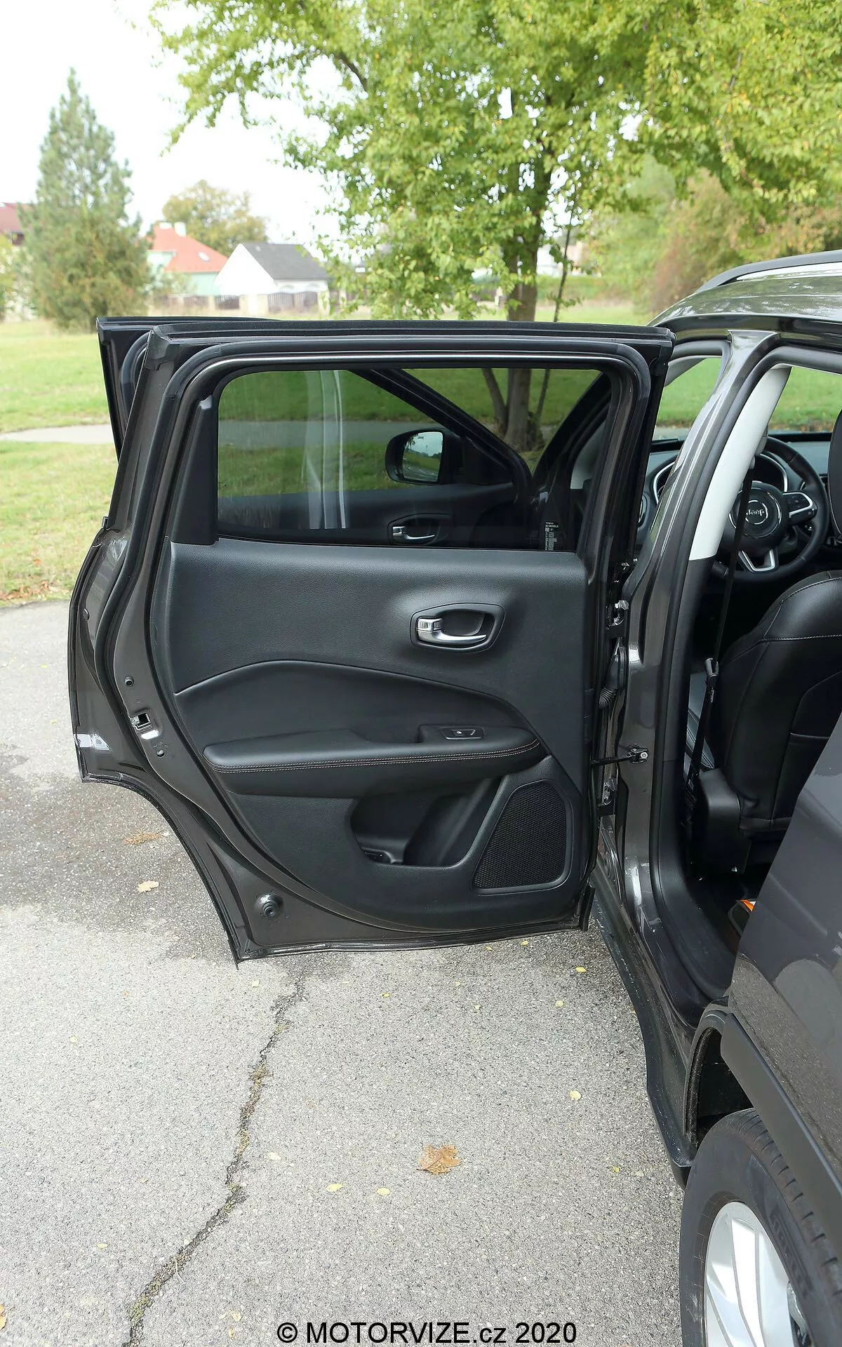 Vue intérieure d'une Jeep Compass 2017 depuis l'extérieur montrant la porte du conducteur ouverte avec une poignée métallique et ergonomique, des compartiments de rangement, une grille de haut-parleur et un panneau de commande pour les opérations de fenêtre et de verrouillage des portes. Une partie du tableau de bord, de la console centrale et du volant multifonction sont visibles, suggérant un intérieur moderne et riche en fonctionnalités. Lumière naturelle, temps couvert, arbres en arrière-plan suggérant un cadre extérieur dans un parc ou une rue bordée d'arbres.