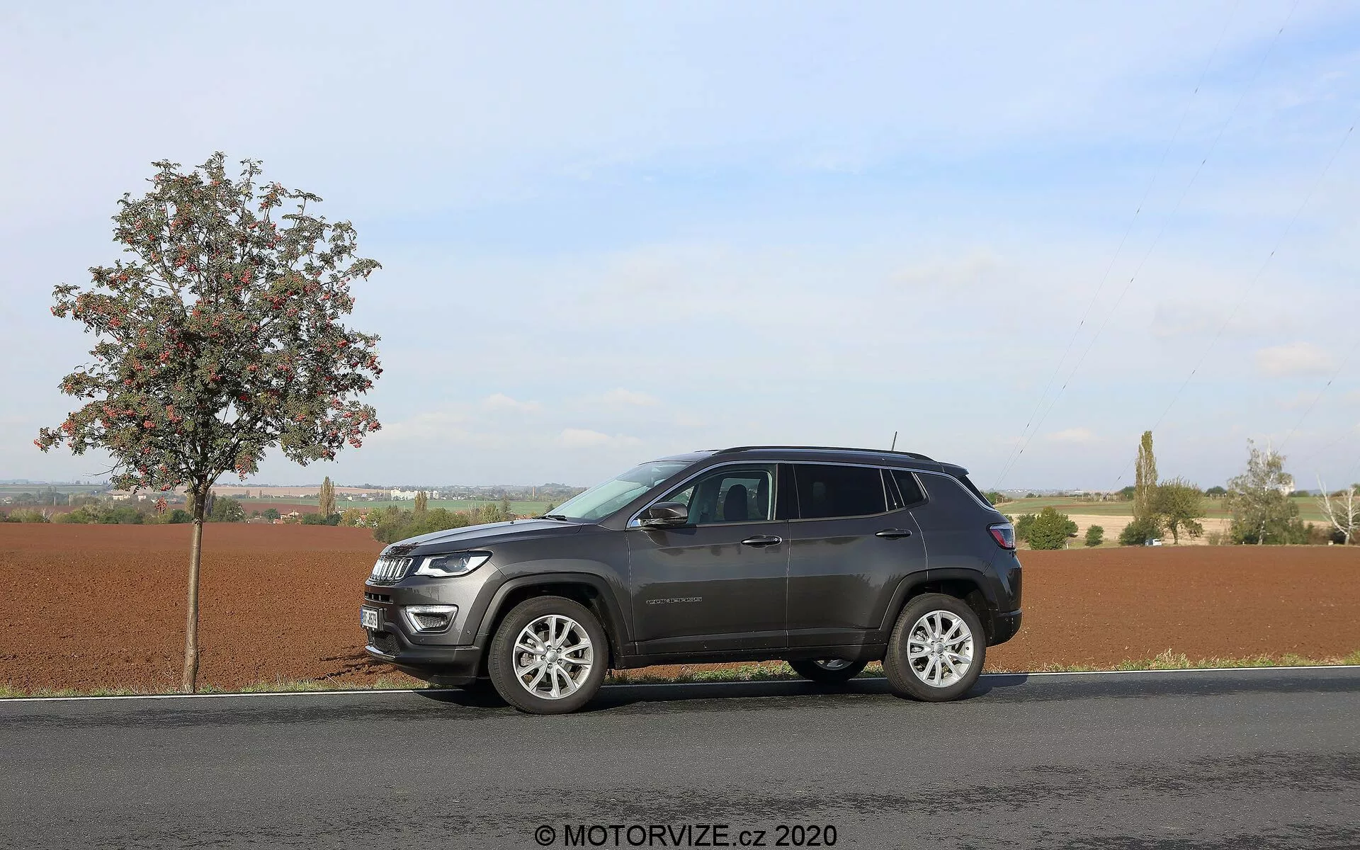 Image d'un SUV Jeep Compass 2017 garé sur une route asphaltée plate à côté d'un jeune arbre feuillu sous un ciel clair et ensoleillé. L'angle latéral capture le profil du véhicule du pare-chocs avant au pare-chocs arrière, mettant en évidence ses éléments de design moderne tels que la grille avant proéminente, les phares élégants, les jantes en alliage multi-branches, les rétroviseurs latéraux prolongés avec des indicateurs de direction possibles et les vitres teintées. Le SUV présente un schéma de couleurs bicolore et un porte-bagages, avec des reflets indiquant des accents chromés sur la façade avant. L'arrière-plan rural suggère une polyvalence pour différents terrains. Filigrane 'MOTORVIZE.cz 2020'.