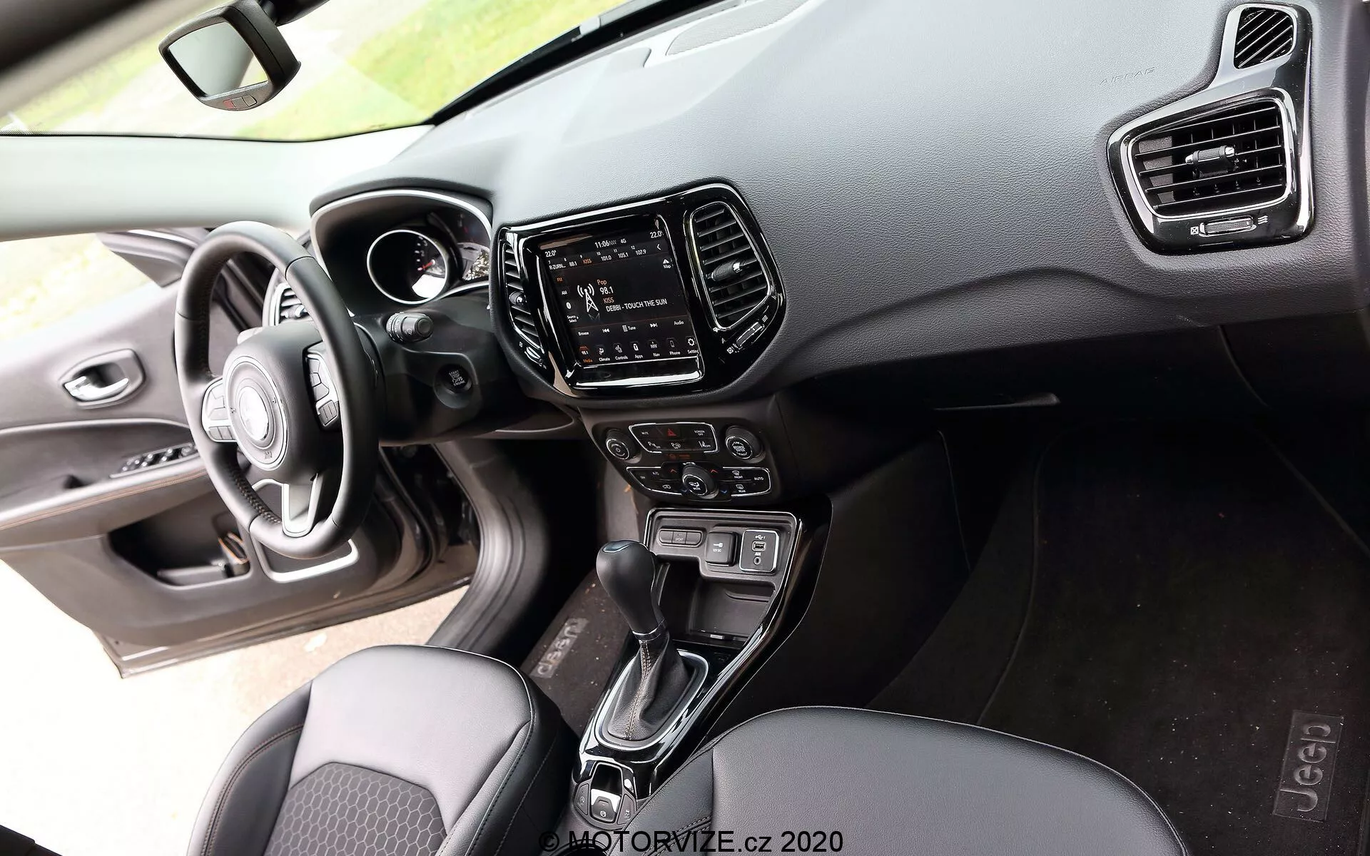 Vue intérieure d'une Jeep Compass 2017 depuis la perspective du passager, montrant un volant à trois branches avec des boutons de commande intégrés, un tableau de bord numérique partiellement visible en matériaux souples au toucher et en plastique dur avec coutures visibles, un système d'infodivertissement à écran tactile dans la console centrale encadré par des évents de climatisation verticaux, un levier de vitesses automatique entre les sièges avant, des boutons et des boutons pour le contrôle du climat, des sièges en tissu avec un motif et un aperçu du panneau de porte du conducteur avec des commandes de réglage des fenêtres et des rétroviseurs latéraux rehaussées par un éclairage d'ambiance, mettant en évidence l'accent sur le confort et la technologie sur le marché de milieu à haut de gamme.
