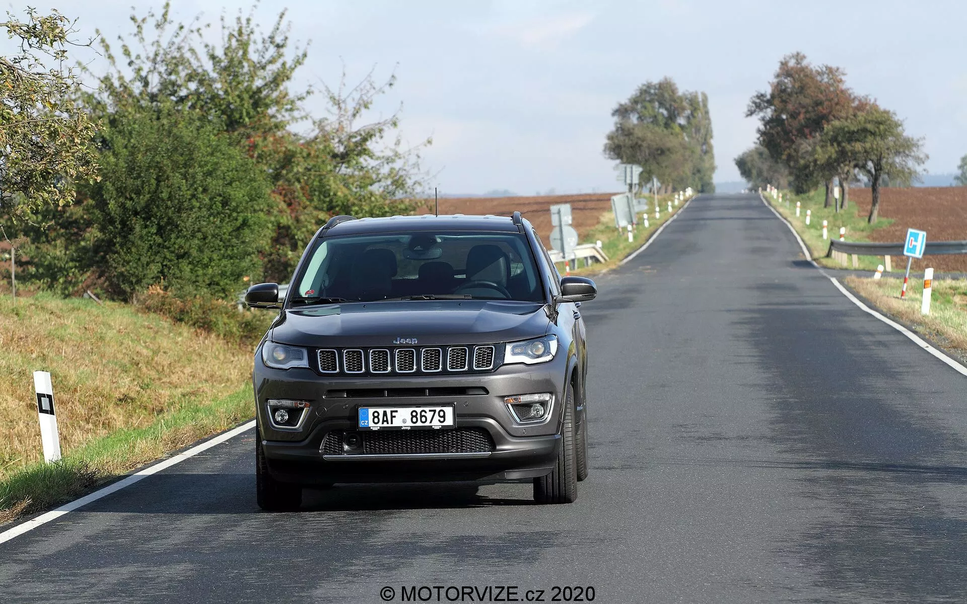 Vue avant d'une Jeep Compass 2017 roulant à vitesse modérée sur une route de campagne. L'image capture les caractéristiques frontales distinctives, y compris la grille avant iconique à sept fentes, les phares rectangulaires modernes et les feux de brouillard intégrés dans le pare-chocs. La voiture est photographiée à hauteur des yeux, mettant en valeur son design extérieur et la technologie d'éclairage sous une bonne lumière naturelle diurne. Une plaque d'immatriculation de style européen est montée au centre à l'avant, avec un paysage champêtre clair en arrière-plan.