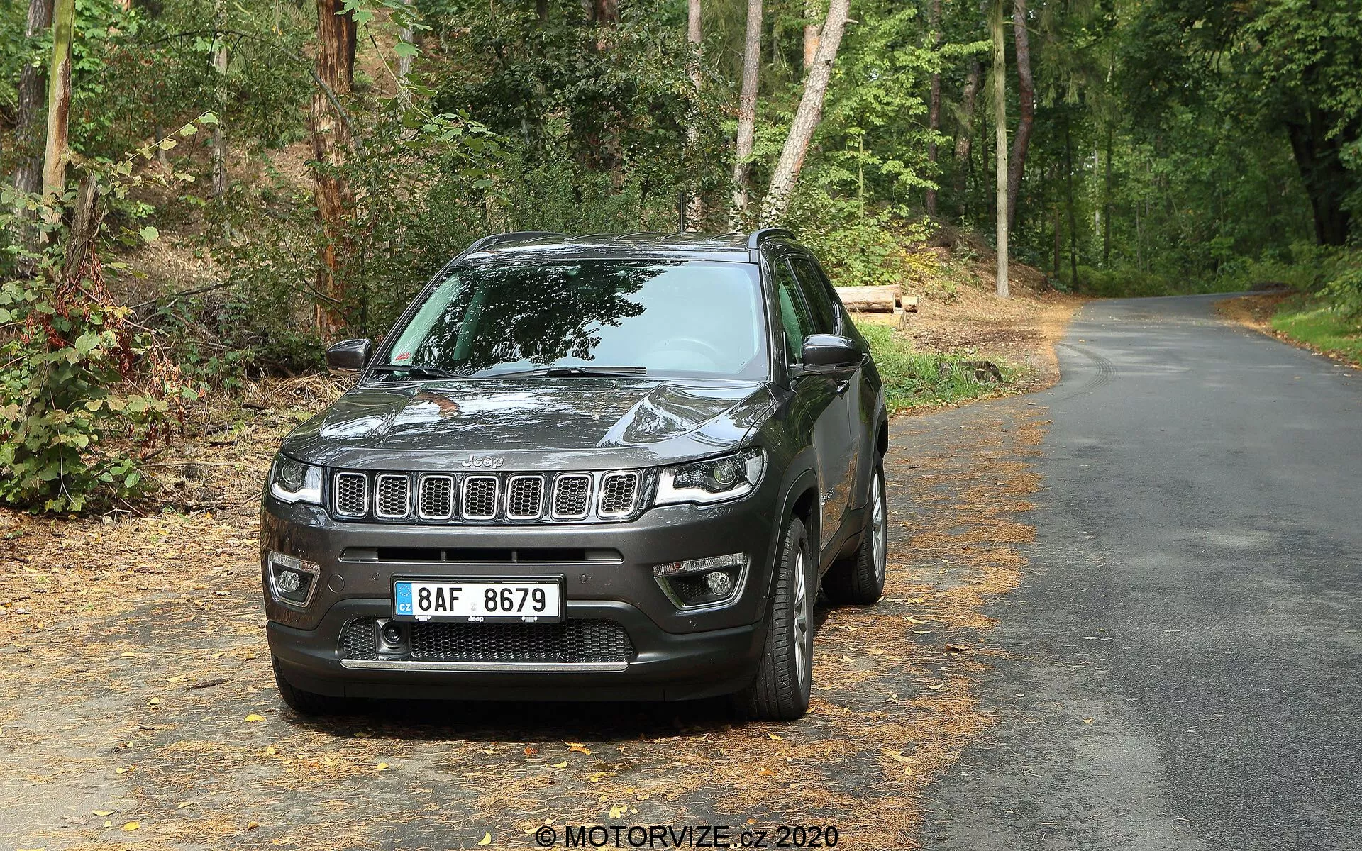 SUV Jeep Compass 2017 sur une route pavée entourée de verdure, faisant face à la caméra sous un angle légèrement à gauche montrant l'avant et le côté conducteur. Les caractéristiques visibles comprennent une calandre avant à barres horizontales avec un emblème centré, des phares rectangulaires à LED, des logements pour phares antibrouillard dans le pare-chocs inférieur, un capot contouré reflétant la lumière, des capuchons de rétroviseurs de la couleur de la carrosserie, des fenêtres légèrement teintées, une roue en alliage à plusieurs rayons et une aile avant profilée. L'arrière-plan comporte des arbres et du feuillage indiquant une zone routière boisée.