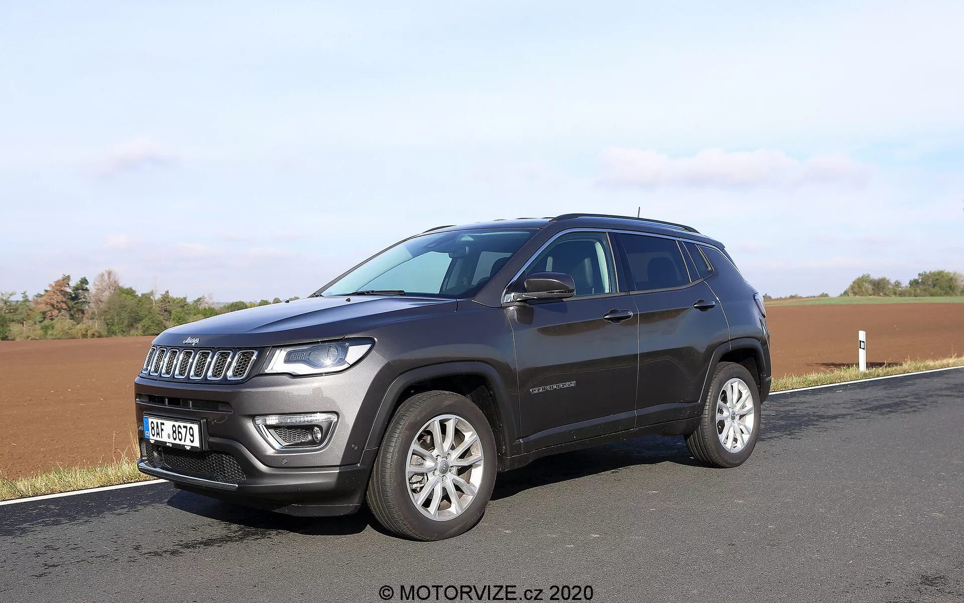 SUV moderne garé le long d'une route avec un champ en arrière-plan, capturé sous un angle avant trois quarts. La photo met en avant la calandre avant à sept fentes du SUV, les phares distinctifs équipés de LED, les jantes en alliage et la peinture métallique foncée. Les détails latéraux tels que la ligne de fenêtre, les poignées de porte, les rétroviseurs latéraux éventuellement avec clignotants intégrés et une ligne de toit légèrement inclinée sont visibles. Un badge sur la porte avant est également visible, fournissant des informations potentielles sur le niveau de finition.