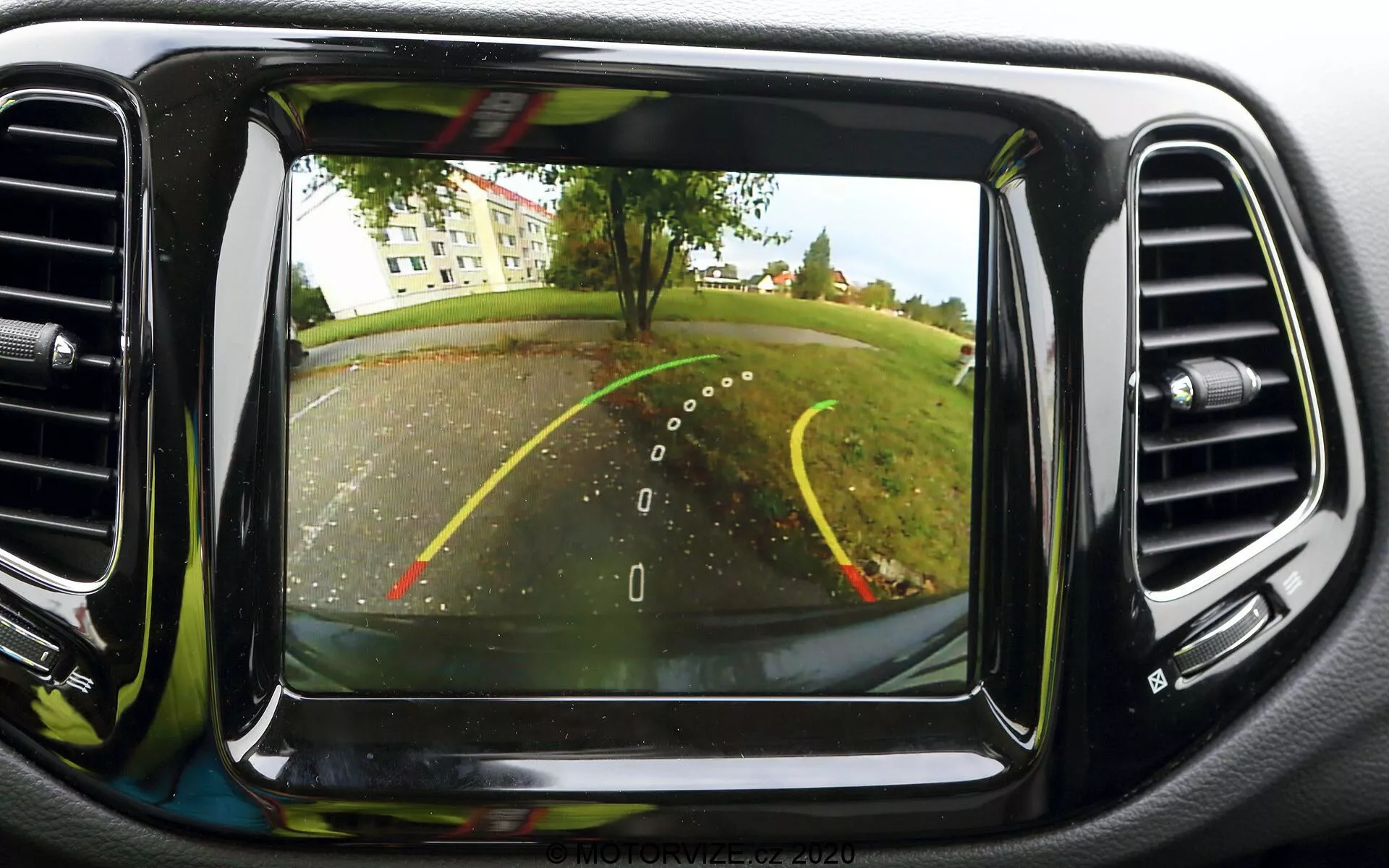 Image du système de caméra de recul sur l'écran de tableau de bord d'une Jeep Compass 2017. La caméra, située à l'arrière près de la plaque d'immatriculation, offre une vue grand angle derrière le véhicule. Les lignes de guidage pour la marche arrière sont visibles, indiquant la largeur de la voiture et la trajectoire projetée avec des lignes jaunes et rouges pour la distance. La scène montre un parking ou une allée avec des arbres et une pelouse en arrière-plan. L'écran est placé au centre du tableau de bord, entouré de bouches d'aération de climatisation, et présente une qualité d'image claire, typique des technologies automobiles modernes.