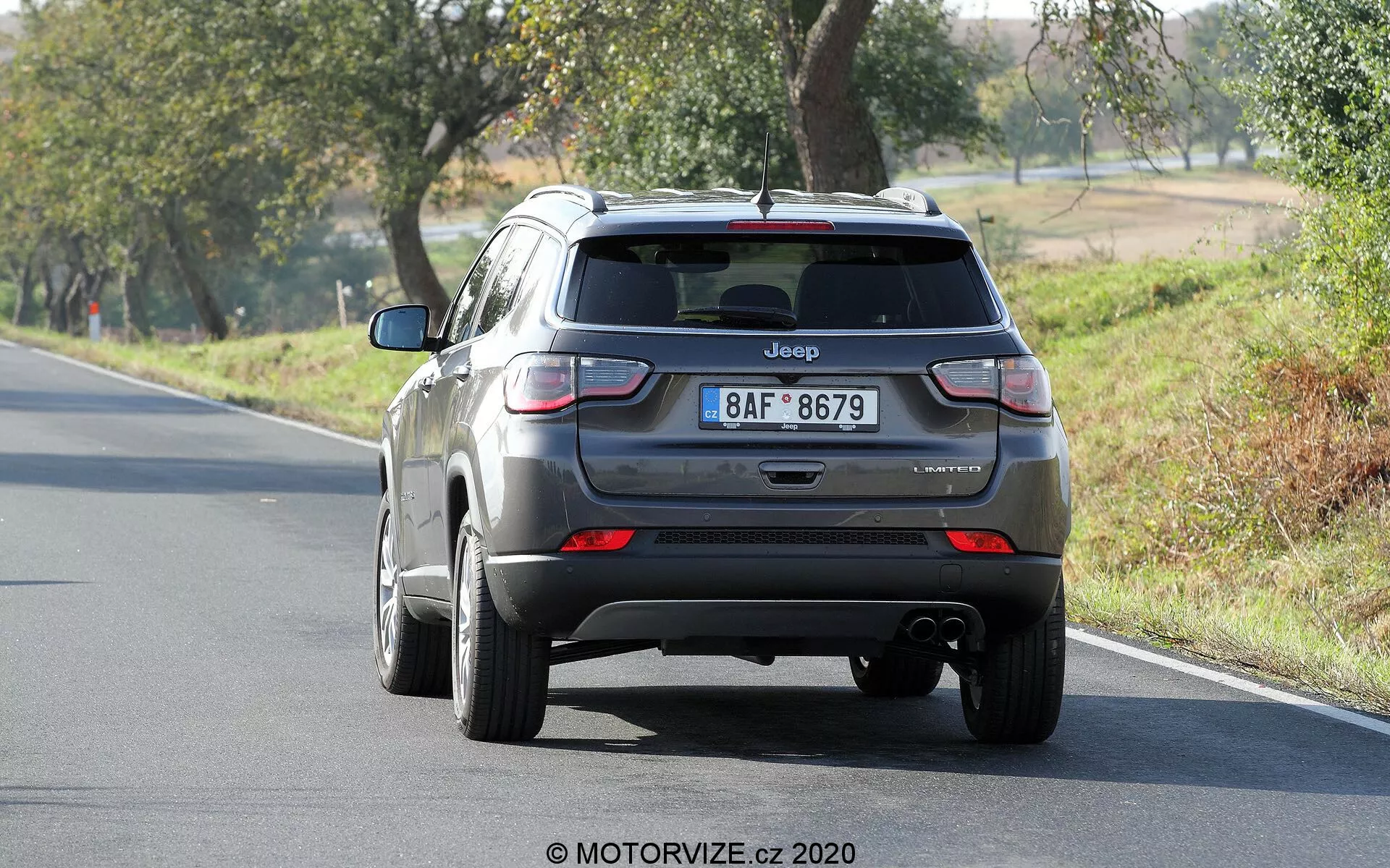 Vue arrière d'une Jeep Compass argentée 2017 présentant le hayon, le pare-chocs arrière et les feux arrière. Le logo 'Jeep' et le nom du modèle 'Compass' sont visibles, avec un badge de finition légèrement flou. L'image montre clairement les feux arrière, l'embout d'échappement sous le pare-chocs et la voiture positionnée sur une route à deux voies entourée d'arbres dans un cadre rural.