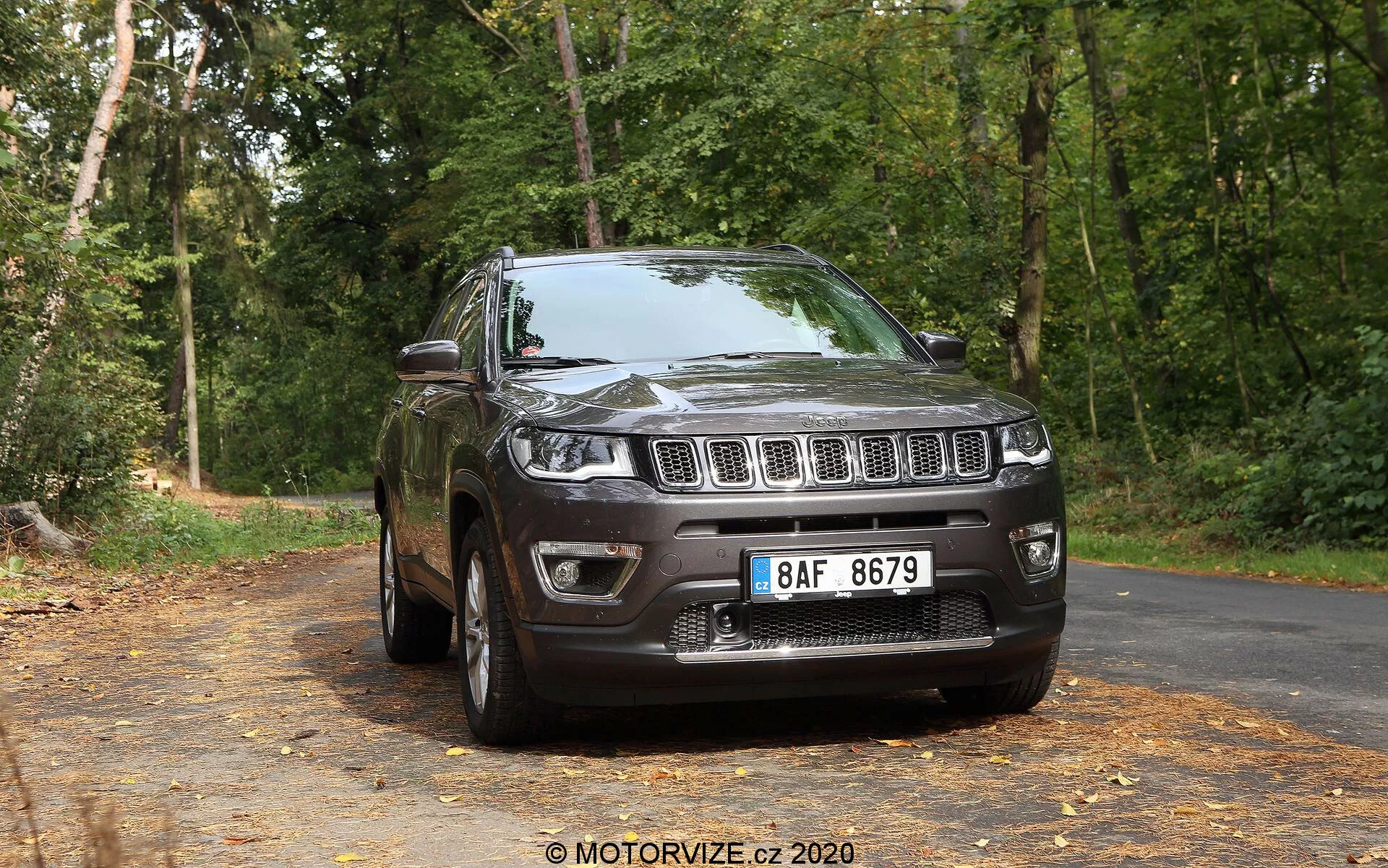 Vue de trois quarts avant d'un SUV Jeep Compass 2017 de couleur sombre sur une route pavée recouverte de feuilles, avec en arrière-plan des arbres dans un cadre forestier. L'image montre la calandre avant avec des fentes verticales distinctives, encadrée par des phares s'étendant dans les ailes, un pare-chocs avant bien défini avec des phares antibrouillard et un capot surélevé sculpté. Le profil latéral révèle des jantes en alliage, une aile visible, des rétroviseurs latéraux, des poignées de porte, des contours de fenêtres et un système de rails de toit, le tout mis en lumière par un éclairage naturel.