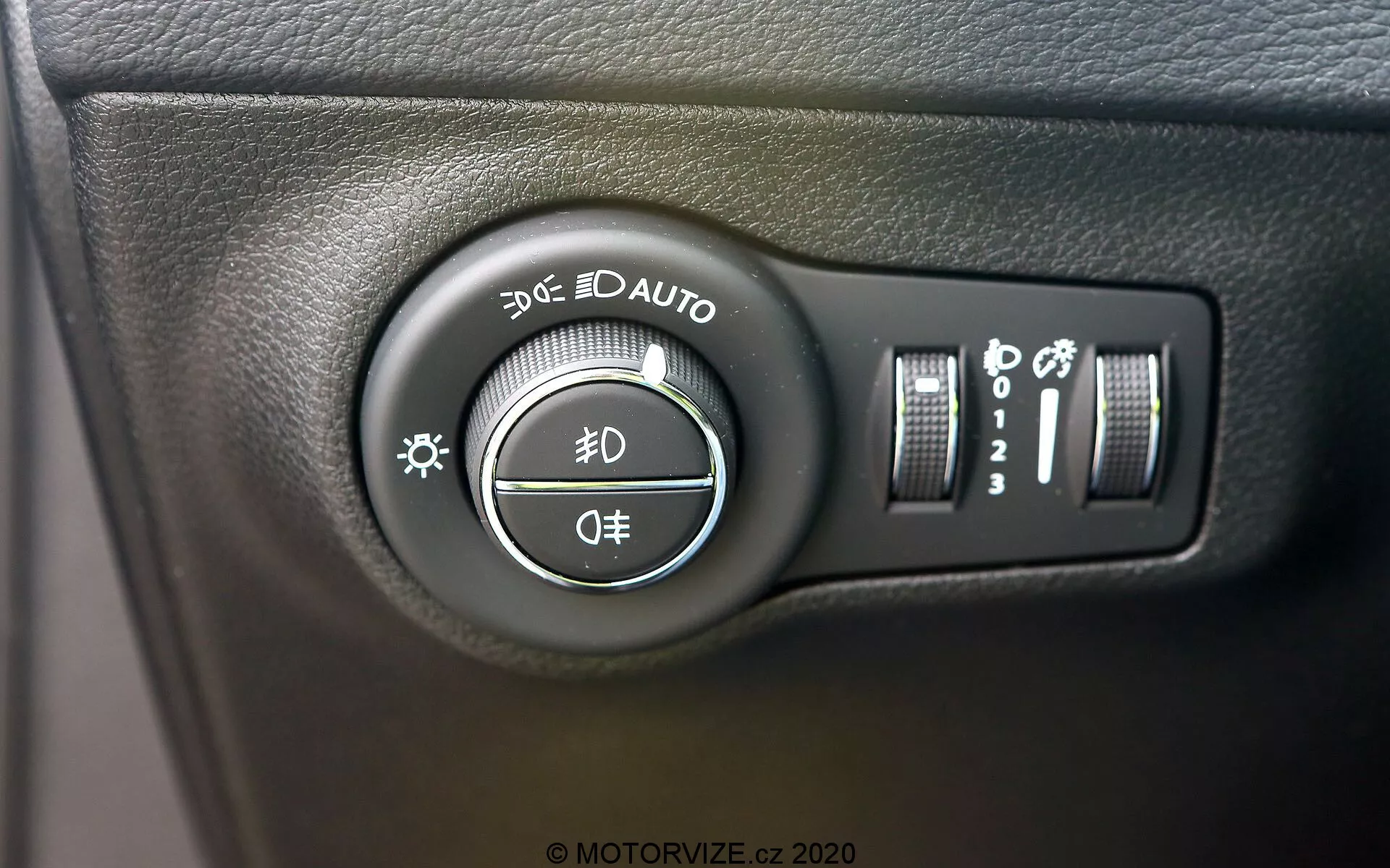 Vue en gros plan de l'interrupteur de commande des phares de la Jeep Compass 2017 situé sur le tableau de bord à gauche du volant. L'image, prise sous un léger angle, met en évidence le sélecteur rotatif avec des réglages pour les feux de position, les feux diurnes, les phares automatiques, les feux de croisement et de route, et une position d'arrêt. Les commandes supplémentaires à droite comprennent un curseur vertical pour la luminosité du tableau de bord et un bouton pouvant indiquer des feux de route automatiques ou un nivellement des phares. L'interrupteur est monté sur une surface en plastique noir texturé, avec des marques blanches claires et des reflets légèrement brillants.