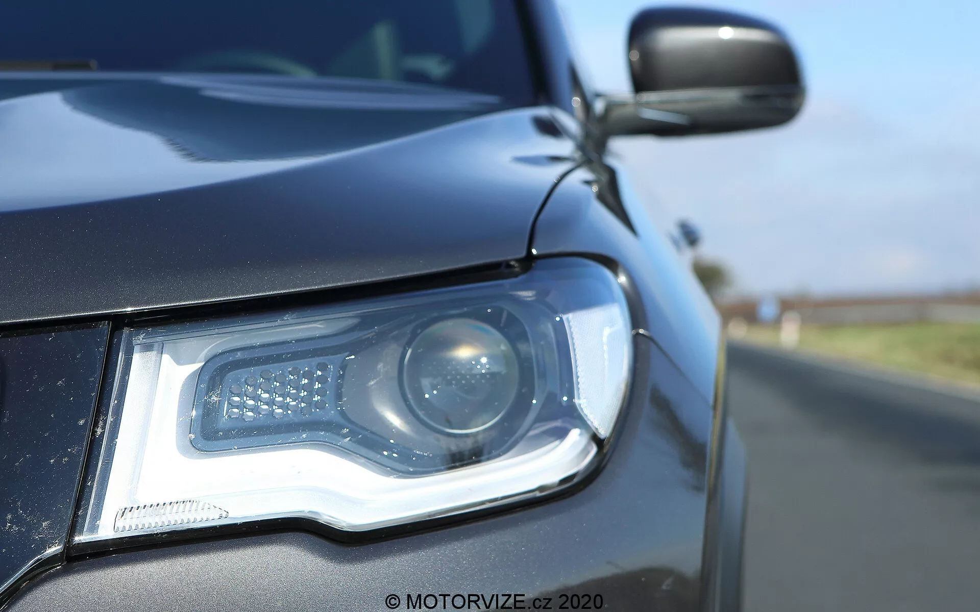 Vue en gros plan du côté avant gauche d'une Jeep Compass 2017, mettant en avant le bloc optique depuis un angle bas à hauteur du sol. Le phare moderne comporte un faisceau de projecteur et des feux de jour à LED disposés dans un motif distinctif. Le boîtier du phare, élégant, s'intègre harmonieusement à la carrosserie de couleur sombre, reflétant un ciel bleu clair. Le rétroviseur latéral, partiellement visible, semble correspondre à la couleur de la carrosserie et peut inclure un indicateur latéral. La faible profondeur de champ met en évidence les détails du phare et de la calandre avant sur un fond flou.