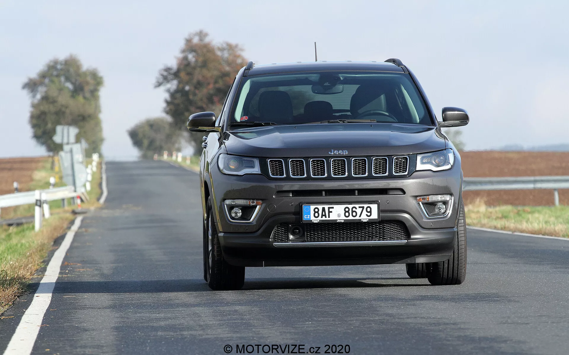 Vue frontale d'un Jeep Compass 2017 en mouvement sur une route à deux voies, capturée sous un angle bas, mettant en évidence la grille à sept fentes proéminente, les phares carrés avec des feux de jour modernes et les feux antibrouillard visibles et les capteurs de stationnement avant sur le pare-chocs. La route s'étend jusqu'à l'horizon avec une double ligne jaune continue, indiquant une zone de non-dépassement. Le flou de l'arrière-plan en mouvement indique que le véhicule est en mouvement, montrant sa posture élevée typique des SUV.