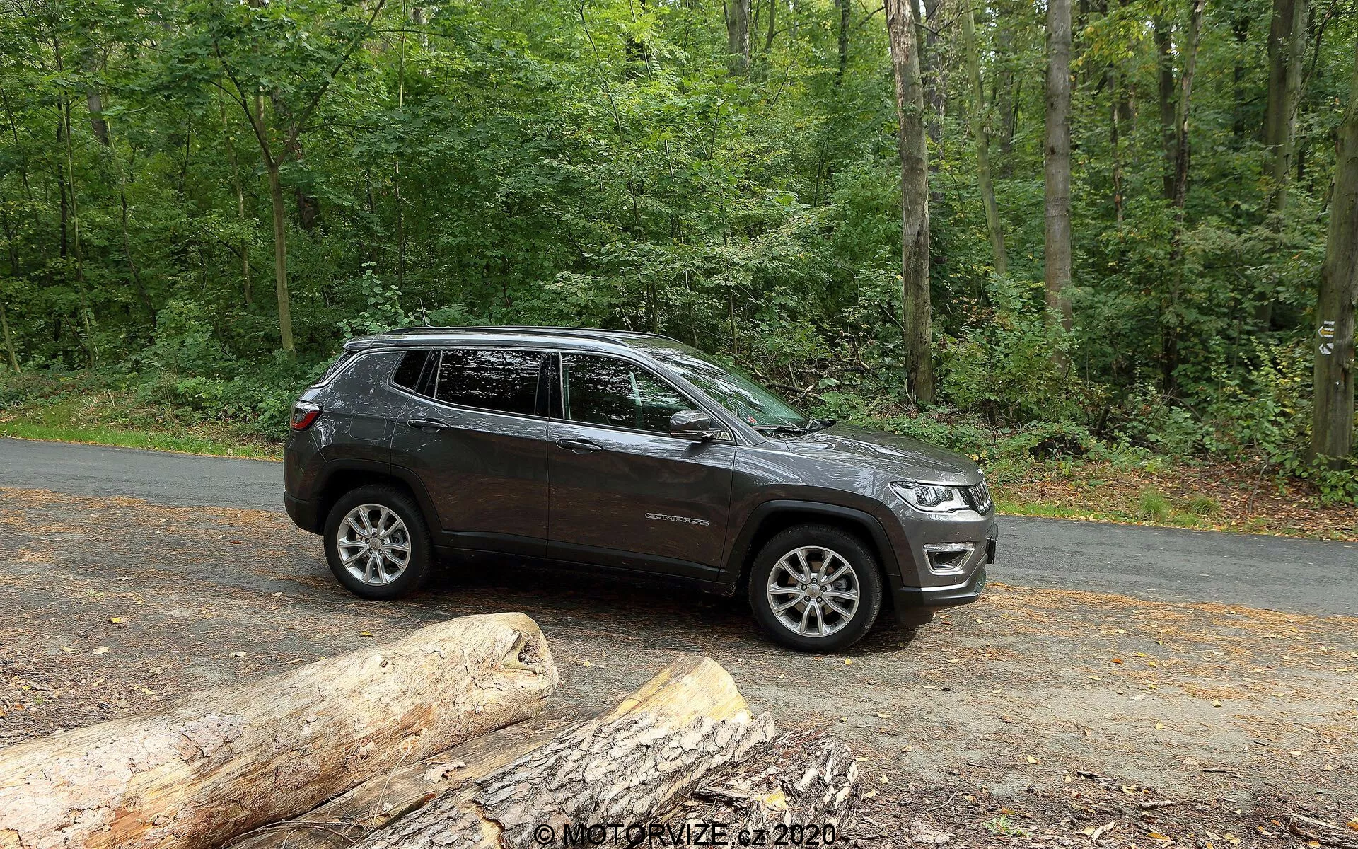 Vue latérale d'un SUV Jeep Compass 2017 stationné sur une route asphaltée dans un cadre naturel avec des arbres verts luxuriants. Le véhicule présente des lignes de design modernes le long des portes et des arches de roue, avec des roues en alliage visibles au design multi-rayons. Il a une garde au sol élevée suggérant des capacités tout-terrain. L'image capture une partie de la calandre avant et des phares, indiquant un style contemporain. Des rails de toit, des clignotants intégrés dans les rétroviseurs latéraux et des vitres teintées sont présents. La peinture de couleur sombre avec une finition brillante indique un véhicule bien entretenu. À l'arrêt, le SUV est montré avec une lumière naturelle abondante, idéal pour montrer ses caractéristiques et son attrait pour les amateurs de plein air et d'aventure.