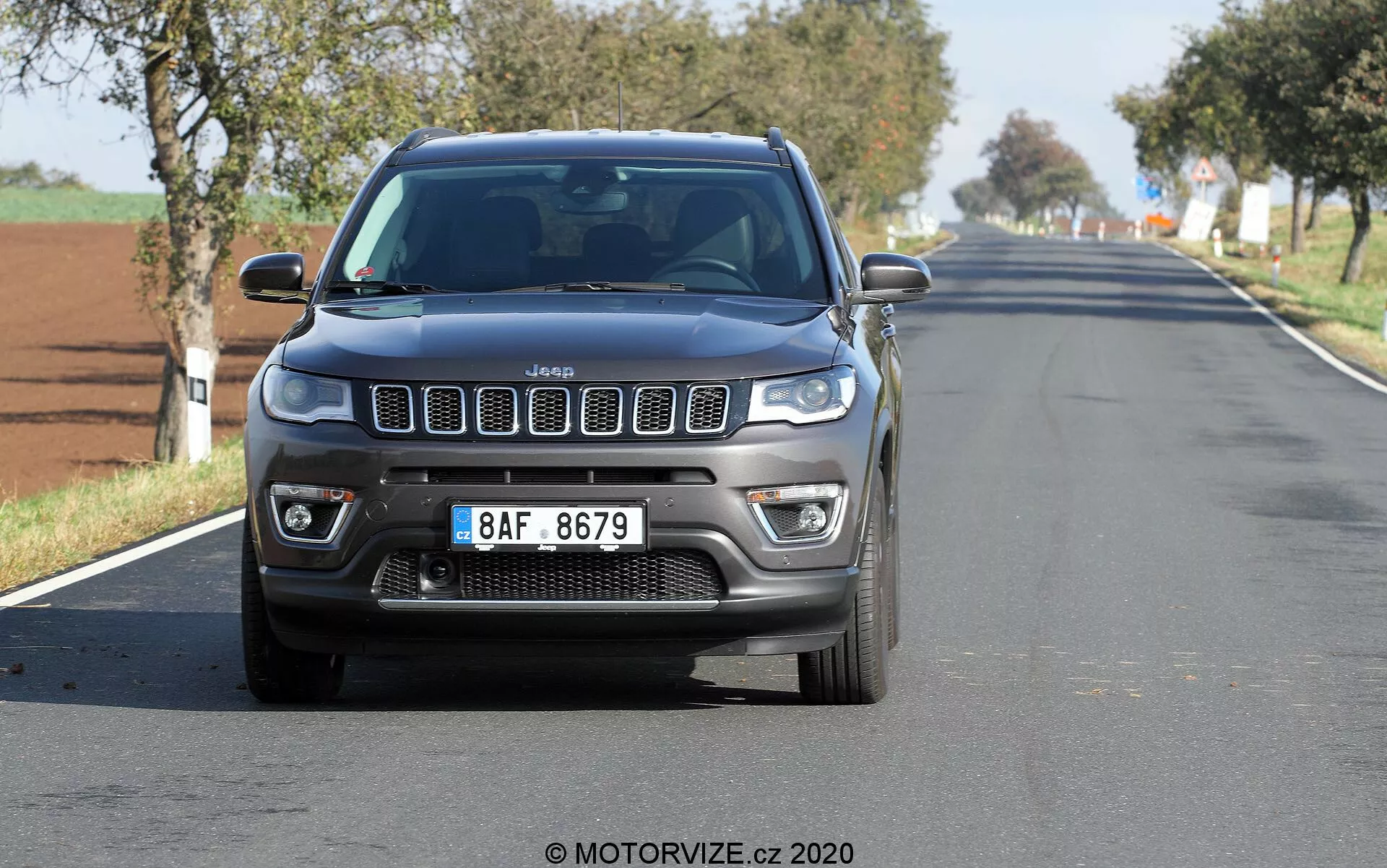 Vue frontale d'une Jeep Compass 2017 circulant sur une route rurale ou suburbaine à deux voies, capturée sous un angle bas, mettant en avant sa calandre iconique à sept fentes, feux de jour à LED, phares antibrouillard et contours robustes du capot. La voiture est centrée et relativement proche de la caméra, avec un léger flou sur les roues et l'arrière-plan. Le ciel dégagé suggère une journée ensoleillée.