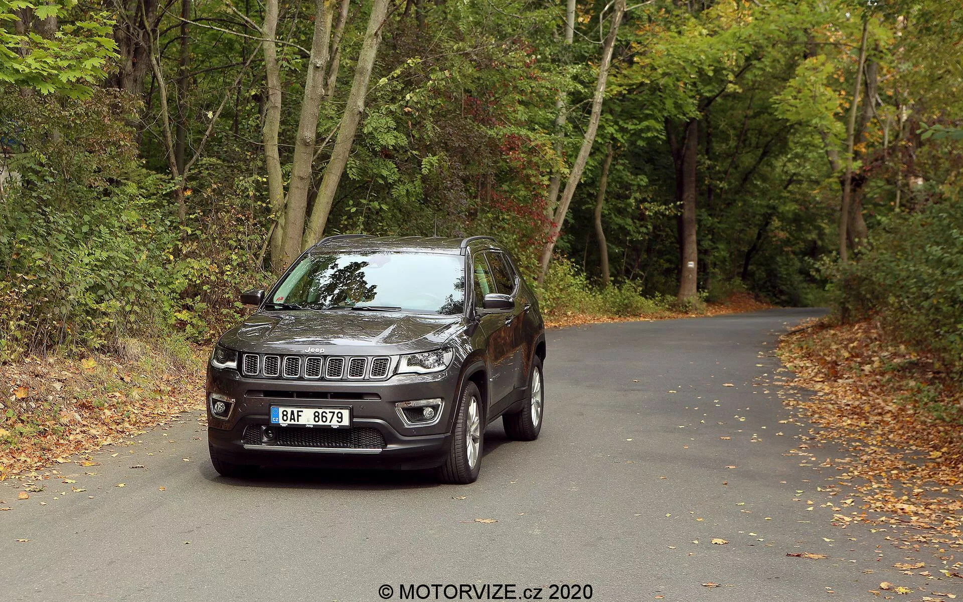 Un SUV Jeep Compass de 2017 positionné sur une route goudronnée entourée de bois en automne. Le véhicule est photographié sous un angle trois quarts frontal, montrant la grille distincte à sept fentes de Jeep et l'emblème central, des phares LED modernes et des phares antibrouillard intégrés dans le pare-chocs. Il est équipé de roues en alliage, de rétroviseurs latéraux avec clignotants intégrés, d'une structure robuste avec des habillages autour des arches de roue et de vitres légèrement teintées. La vue détaillée met en valeur les éléments de design et technologiques, suggérant son aptitude aux aventures urbaines et tout-terrain.