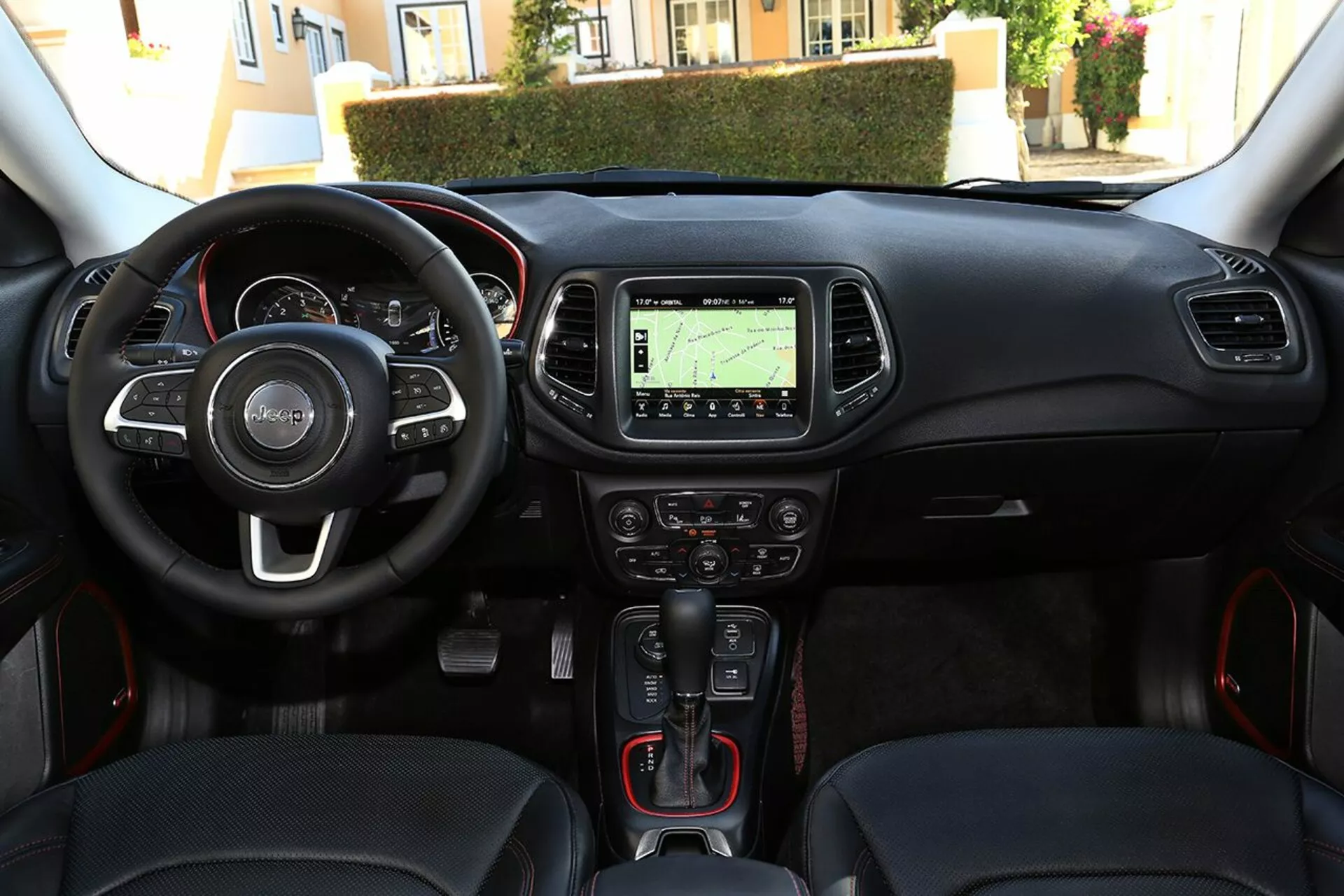 Vue intérieure du Jeep Compass 2017 du point de vue du conducteur, montrant le volant avec commandes intégrées, cluster d'instrumentation, console centrale avec système de navigation, commandes climatiques, levier de changement de vitesse automatique avec détails rouges sportifs, bouton de démarrage-arrêt sans clé, et port de connectivité multimédia