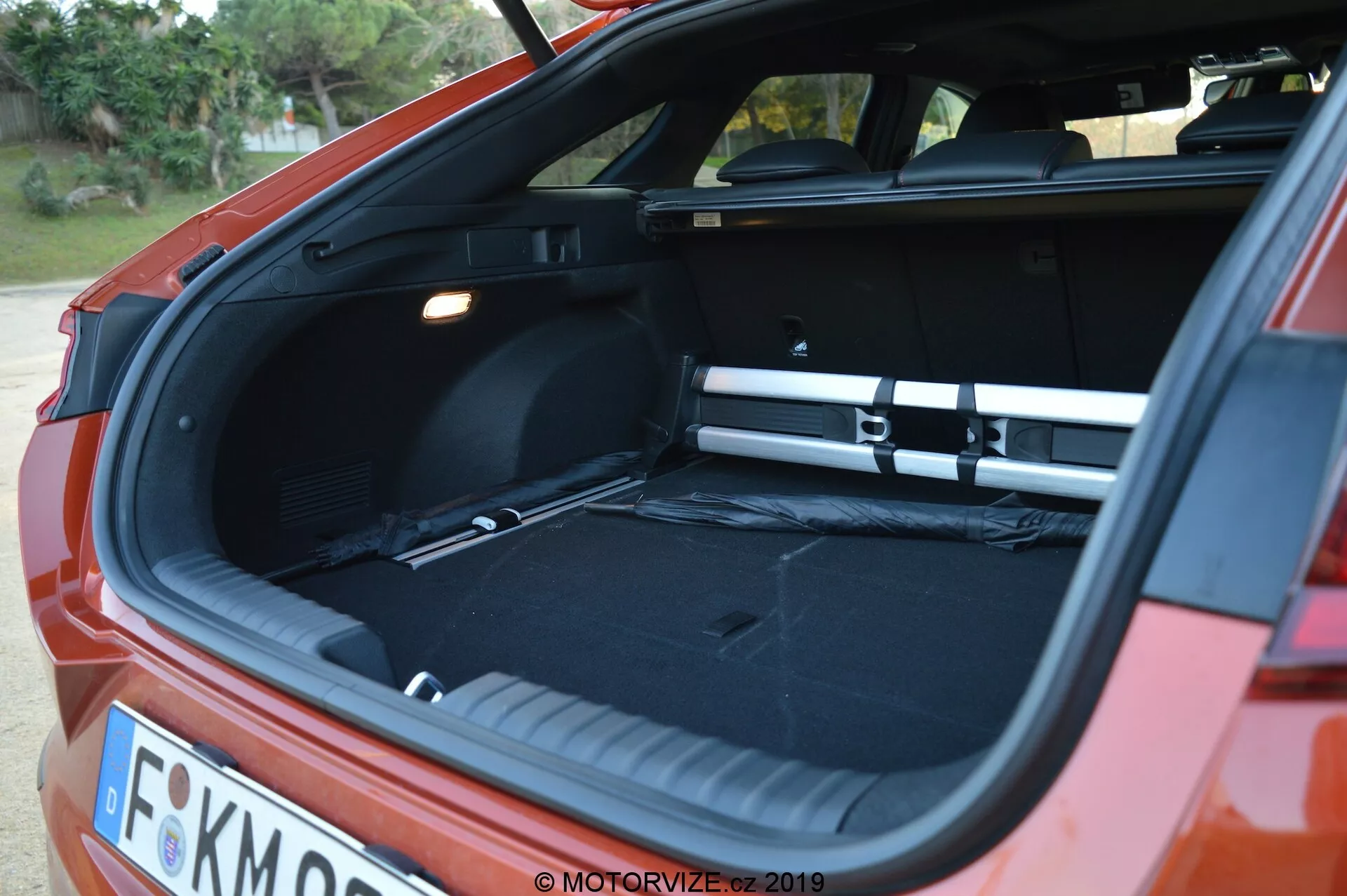 L'image capture une vue arrière angulaire d'une Kia Ceed 2021 rouge, mettant en évidence le coffre à hayon ouvert. Il fait soit l'aube soit le crépuscule, et un éclairage doux met en valeur l'espace de chargement vide et spacieux avec un intérieur noir. Une couverture de chargement est en place, s'étendant sur toute la largeur du coffre. La photo, prise depuis le coin arrière avec un léger angle latéral, montre également des parties du pare-chocs arrière, des feux arrière et une plaque d'immatriculation partiellement visible. Le coffre dispose d'un plancher de chargement plat, d'une source de lumière à gauche et d'un compartiment latéral à droite.
