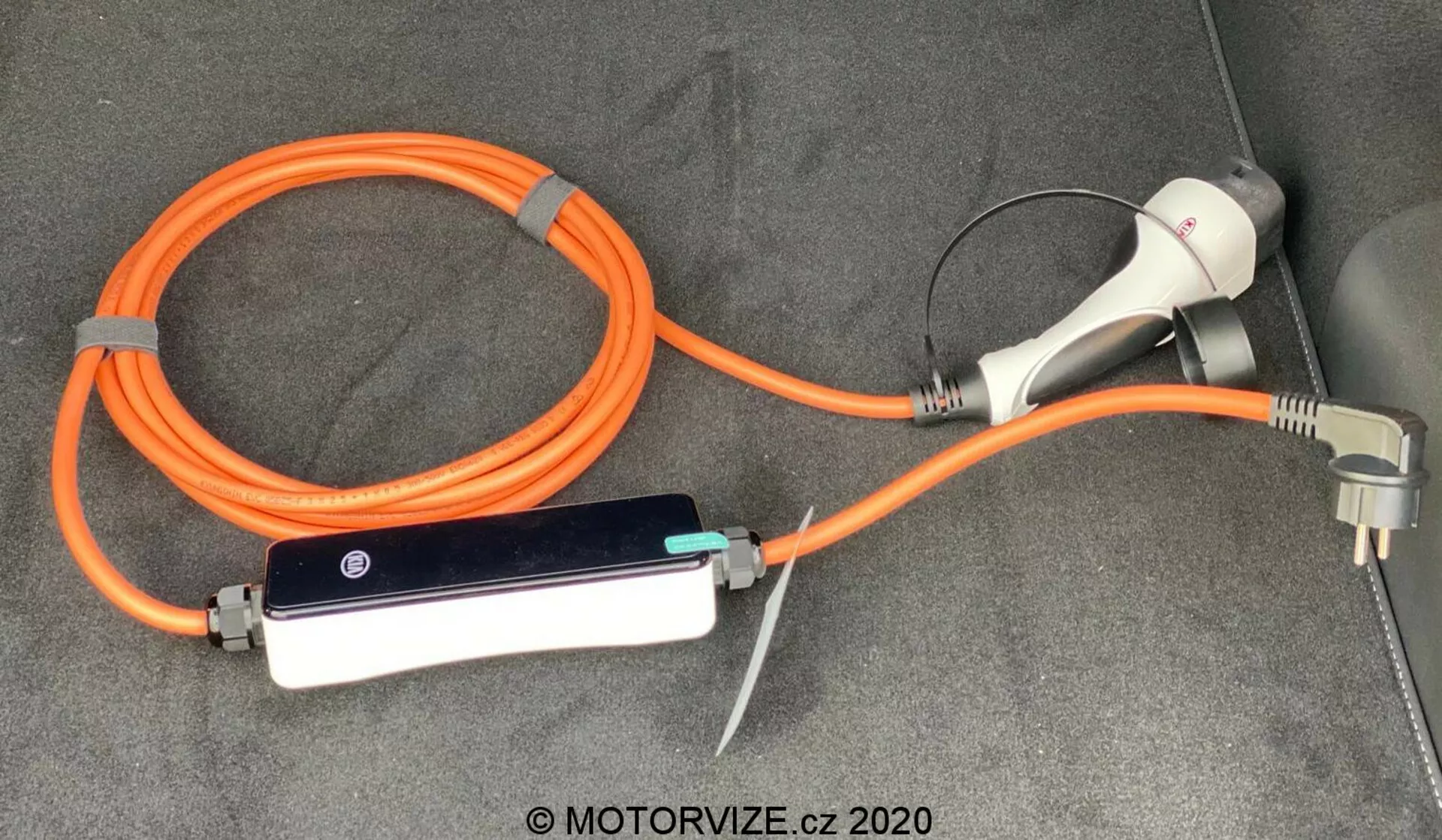 Câble de charge et adaptateur pour véhicule électrique (VE) pour la Kia Ceed 2018, avec un EVSE (Équipement d'Alimentation pour Véhicule Électrique) portable orange à haute visibilité pour la sécurité. L'ensemble comprend un connecteur et une fiche de charge pour une utilisation avec une prise domestique, ainsi qu'un boîtier de contrôle en ligne avec des indicateurs LED, le tout disposé sur le plancher recouvert de moquette du coffre ou de la zone de chargement du véhicule.