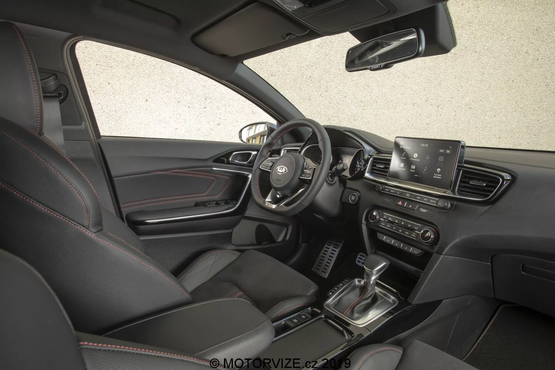 Vue intérieure de la Kia Ceed 2021, montrant le côté conducteur sous un angle haut et légèrement orienté vers l'avant. L'image, capturée depuis le côté passager, offre une vue complète du tableau de bord, y compris le volant central avec boutons de commande, une visibilité partielle du tableau de bord, un écran d'infodivertissement bien en vue, des bouches d'aération, des boutons et des boutons de commande de climatisation, le levier de changement de vitesse, le système de démarrage par bouton-poussoir, des sièges en cuir avec commandes électriques et une palette de couleurs intérieures sombres.