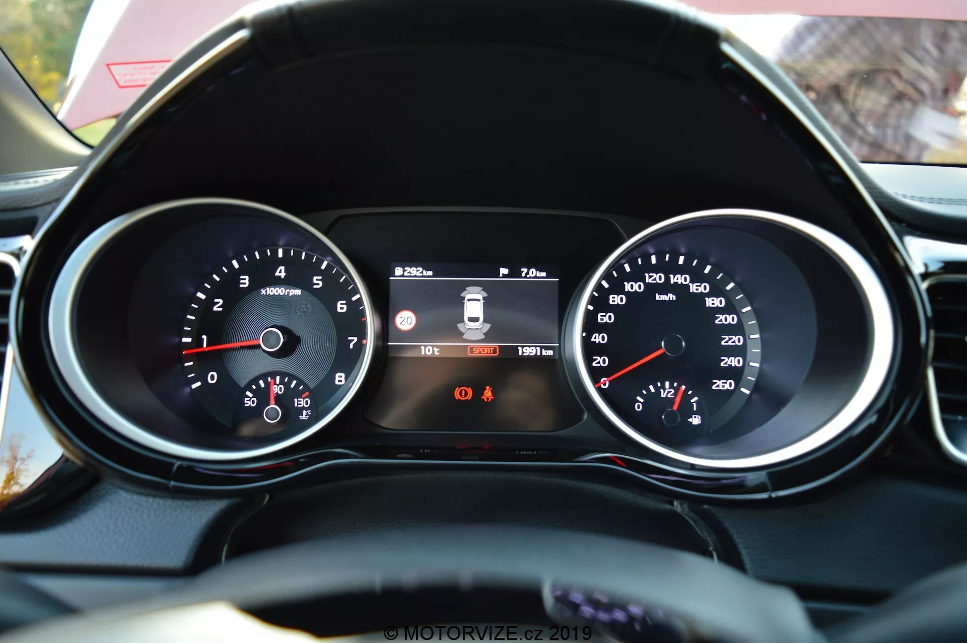Gros plan depuis la perspective du conducteur du tableau de bord de la Kia Ceed 2021 et d'une partie du volant fini en cuir. Les caractéristiques comprennent un tachymètre à gauche avec des lectures en RPM, un affichage numérique central montrant le système d'avertissement de franchissement de ligne, l'heure et le compteur kilométrique, et un compteur de vitesse à droite avec la vitesse en km/h. Sous l'affichage central, deux voyants d'alerte sont visibles, le rappel de ceinture de sécurité étant identifiable.