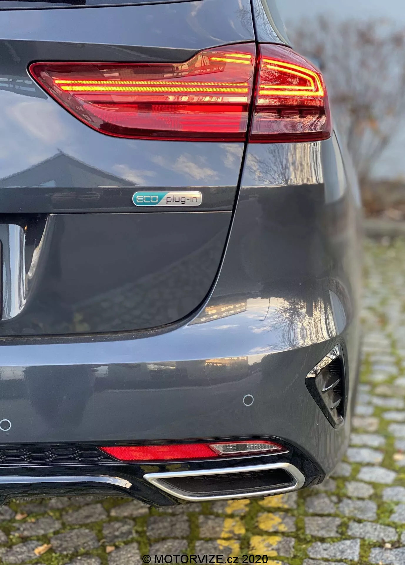 Vue détaillée de l’arrière d’une Kia Ceed 2018 gris métallisé, mettant en évidence le feu arrière LED allumé, le badge eco plug-in, le pare-chocs arrière sculpté avec réflecteur et diffuseur, et les doubles sorties d'échappement chromées. La perspective montre la convergence du pare-chocs arrière et du côté droit, soulignant les éléments des véhicules électriques hybrides et les caractéristiques du design moderne.