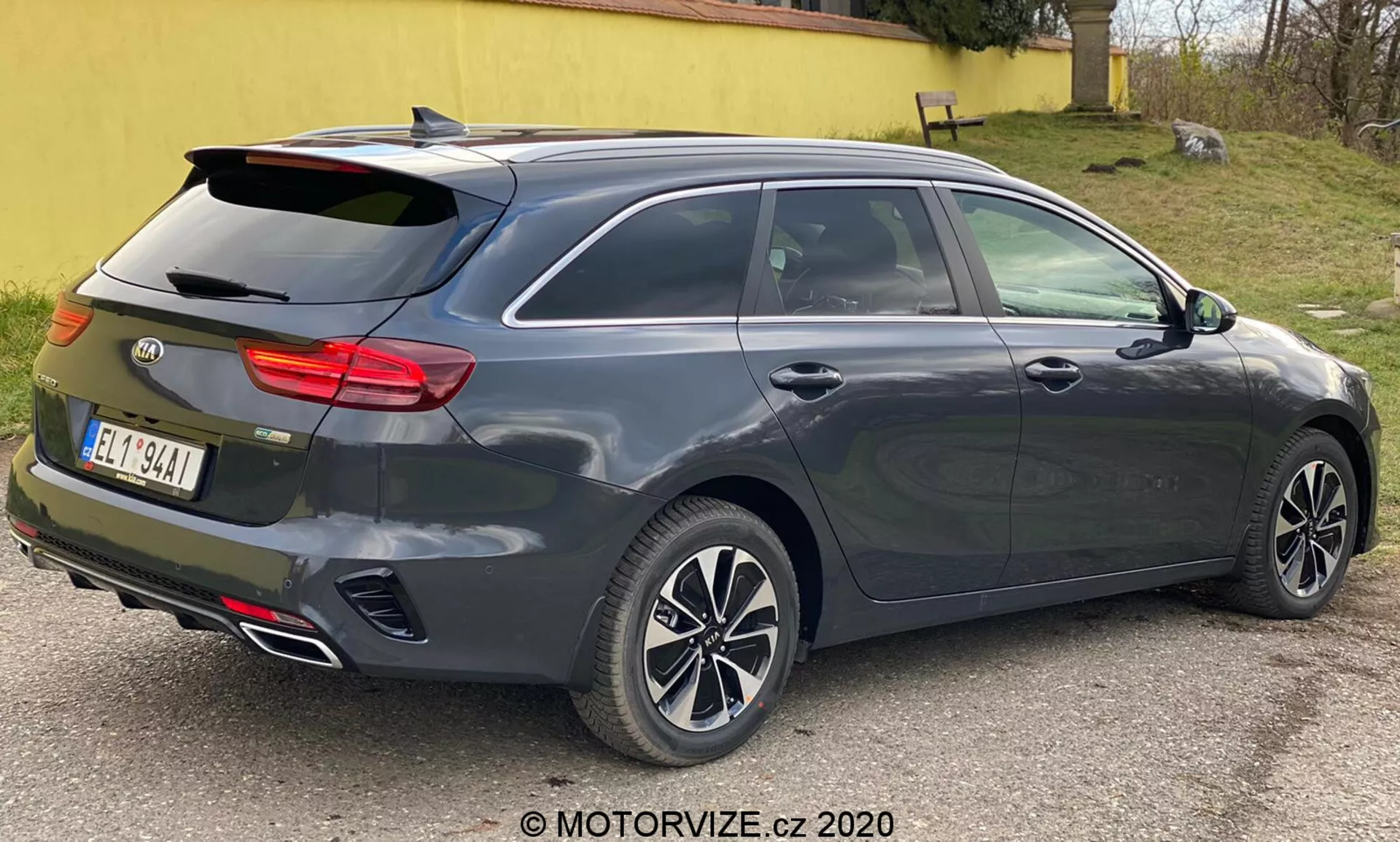 Une Kia Ceed 2018 métallique gris foncé, vue depuis un angle arrière trois-quarts, mettant en avant son design moderne de berline à hayon ou break avec une fenêtre arrière inclinée, spoiler arrière intégré, jantes en alliage à plusieurs branches, poignées de portes et rétroviseurs de couleur carrosserie probablement avec clignotants intégrés, ensemble de feux arrière horizontaux distinctifs peut-être avec technologie LED, antenne aileron de requin, doubles sorties d'échappement, troisième feu stop monté en hauteur, capacité de dégivrage de la fenêtre arrière, capteurs ou réflecteurs indicatifs d'un système d'assistance au stationnement arrière, photographiée de près avec un éclairage neutre.