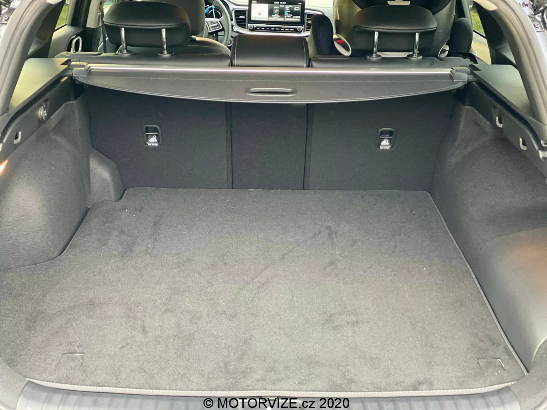 Vue rapprochée du coffre de la Kia Ceed 2018 depuis une perspective arrière, mettant en évidence la zone de chargement spacieuse et propre avec une surface recouverte de moquette. Le coffre est ouvert, révélant un cache-bagages rétractable, les dossiers des sièges arrière de couleur sombre, probablement en cuir, et des languettes argentées pour rabattre les sièges. Les côtés sont doublés d'un matériau doux et un petit compartiment est visible sur le côté droit. Image prise sous un léger angle, montrant la conception et les matériaux de l'intérieur du coffre de la voiture.
