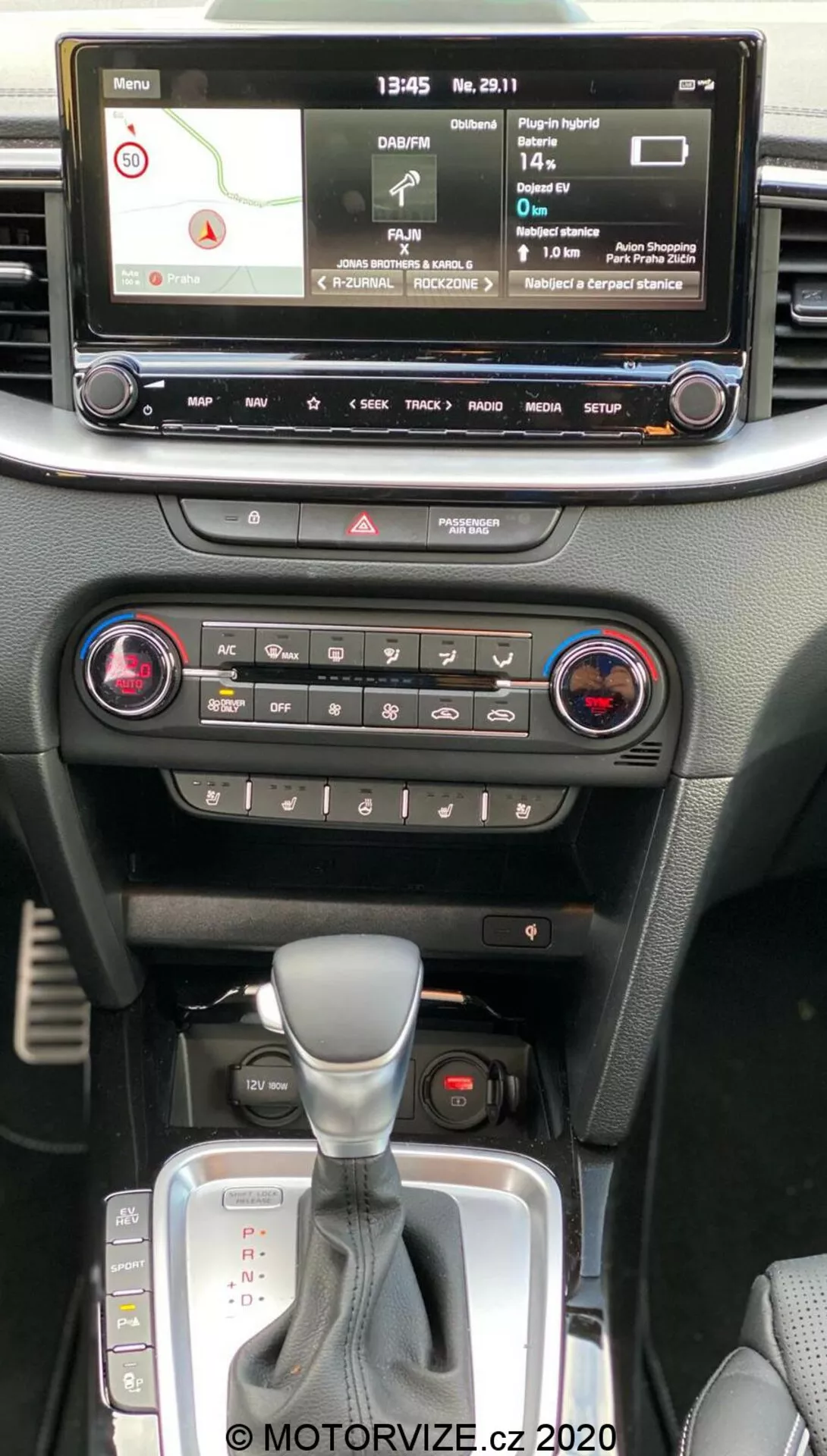 Intérieur de la Kia Ceed 2018 montrant la console centrale depuis la perspective du passager au premier rang, avec écran d'info-divertissement numérique affichant carte de navigation, lecteur multimédia, paramètres du véhicule, commandes de climatisation à double zone, bouton de feux de détresse, levier de changement automatique avec housse en cuir et emplacement pour clé de démarrage sans clé.