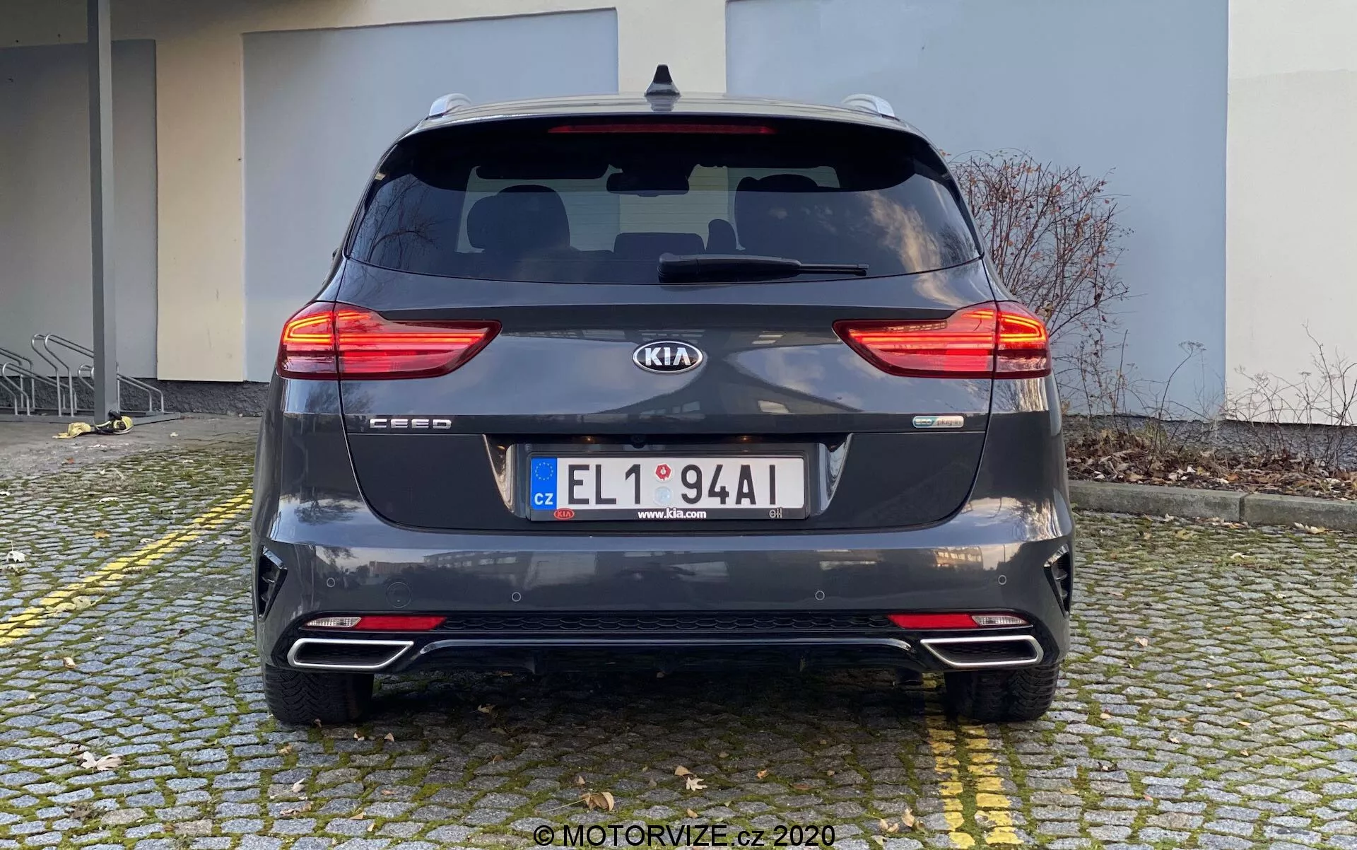 Vue arrière d'une Kia Ceed 2018 garée sur un sol pavé près d'un trottoir. La voiture, capturée en plein jour sous un angle droit à hauteur des yeux, met en valeur ses feux arrières à LED segmentés, lunette arrière avec essuie-glace, emblème du fabricant, nom du modèle, plaque d'immatriculation, pare-chocs arrière avec réflecteurs et capteurs de stationnement, système d'échappement double et finition gris foncé métallisé. L'arrière-plan comprend un bâtiment, indiquant un cadre résidentiel ou commercial.