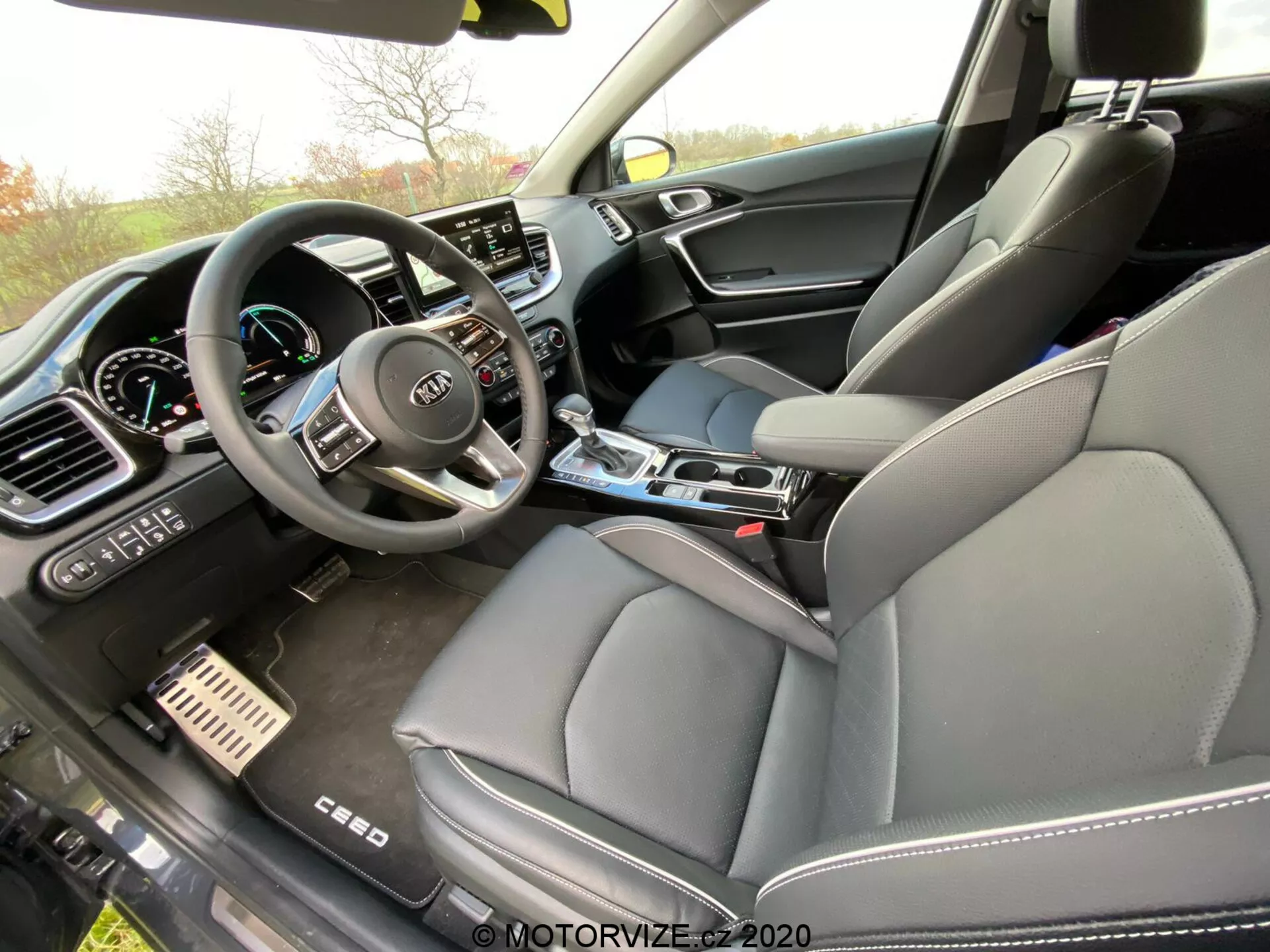 Vue intérieure d'une Kia Ceed 2018 du point de vue du passager, capturant le compartiment avant avec un focus sur le tableau de bord, la console centrale et le siège du conducteur. Le volant avec plusieurs boutons de commande et le logo Kia est visible, ainsi qu'une partie du tableau de bord numérique. La partie centrale du tableau de bord comprend un grand écran tactile d'infodivertissement entouré de réglages manuels de climatisation. La console centrale comprend un levier de vitesse, des porte-gobelets et des cadrans de contrôle. Le siège du passager avant en cuir et le panneau de porte en matériaux mixtes sont également visibles, suggérant une finition luxueuse. Les pédales de frein et d'accélérateur métalliques indiquent également une touche sportive premium.