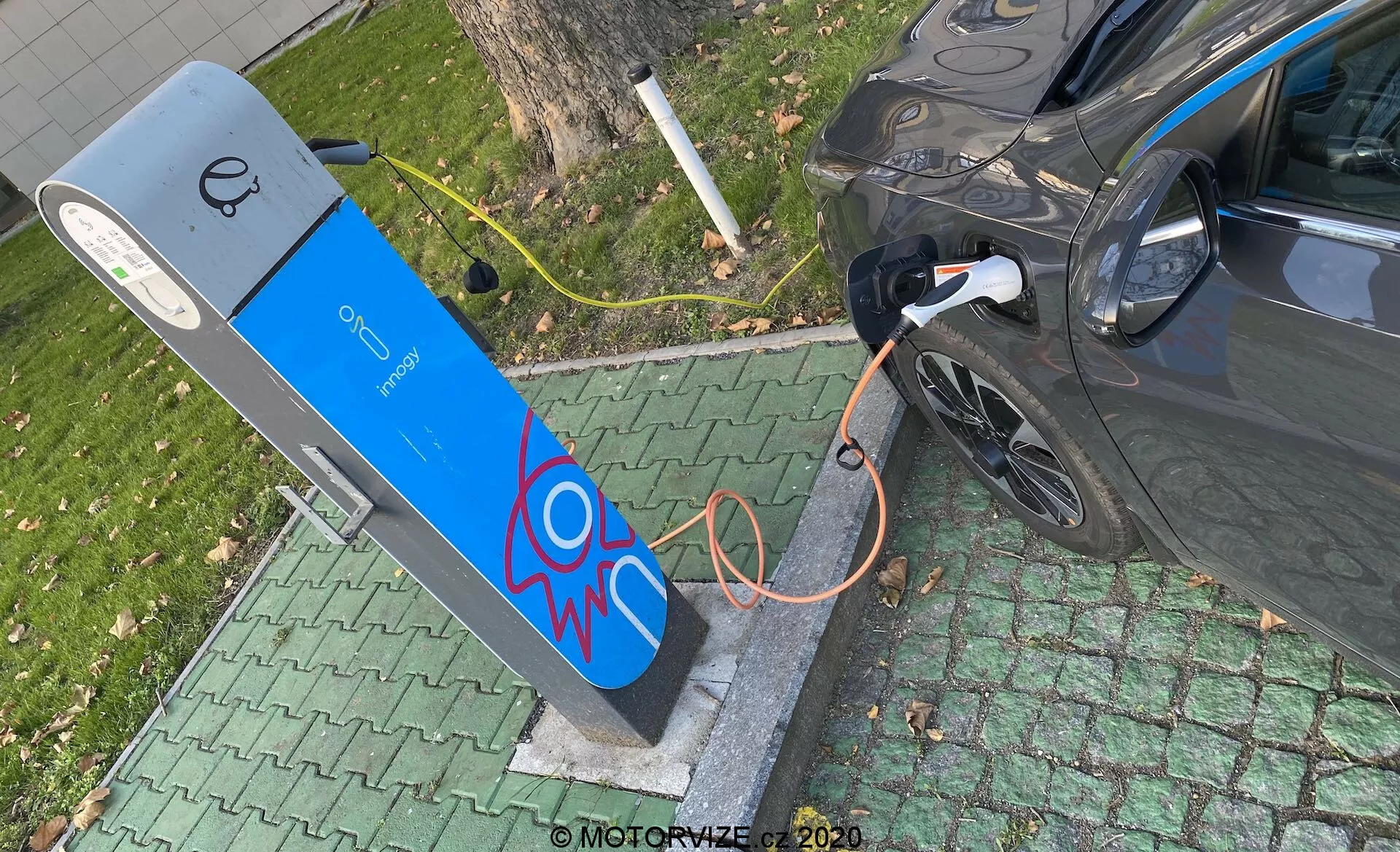 Vue en plongée d'un véhicule électrique moderne en cours de chargement à une borne de recharge publique. L'image montre la section arrière droite de la voiture, en se concentrant sur le port de charge avec le câble orange épais branché. La borne de recharge a une façade bleue avec un graphique de scintillement électrique et un écran en haut, suggérant des éléments d'interface utilisateur. La scène est située dans un environnement vert semblable à un parc, indiquant un emplacement urbain ou suburbain pratique.