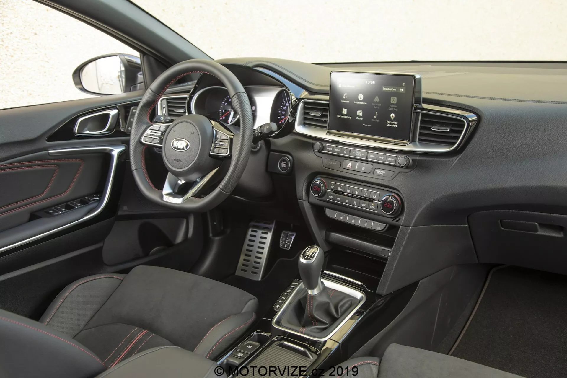 L'intérieur de la Kia Ceed 2021, en se concentrant sur le côté conducteur avec une perspective latérale. Le volant, arborant le logo reconnaissable du fabricant, dispose de commandes multifonctions pour gérer l'infodivertissement, le régulateur de vitesse, la connectivité téléphonique et possiblement les commandes vocales. Derrière, on peut voir le compteur de vitesse, le tachymètre et les jauges d'information du conducteur. La console centrale possède un écran tactile d'infodivertissement offrant navigation, paramètres du véhicule, lecture multimédia et connectivité Bluetooth. En dessous, des commandes manuelles pour les systèmes audio et de climatisation sont présentes. Le levier de vitesses de la transmission manuelle, entouré de boutons pour les modes de conduite ou le contrôle de traction, et les sièges sportifs avec inserts en cuir et tissu, améliorent la sensation premium et orientée performance. Les pédales métalliques dans le repose-pieds du conducteur suggèrent une emphase sur la performance, tandis que les accents chromés sur les bouches de climatisation, les poignées de porte et autres détails ajoutent une touche de sophistication. Le panneau de la porte inclut des commandes pour les fenêtres électriques et possiblement les rétroviseurs électriques, et une partie du rétroviseur est visible, suggérant des fonctionnalités telles que l'assombrissement automatique ou une boussole intégrée.