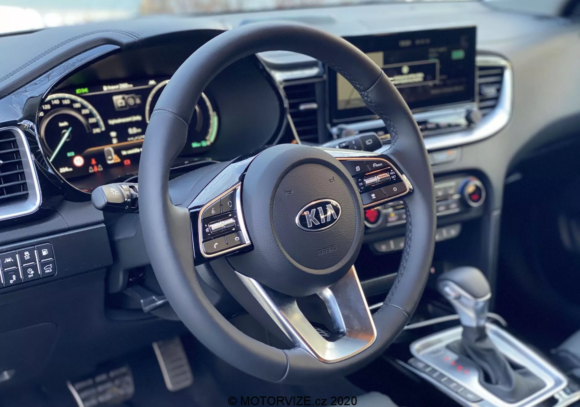 Vue en première personne de l'intérieur de la Kia Ceed 2018 mettant en avant le volant multifonction moderne à trois branches avec logo Kia et le tableau de bord numérique, affichant le compteur de vitesse et le possible compte-tours, ainsi que le système d'infodivertissement à écran tactile, les commandes de climatisation, le levier de changement de vitesse automatique argenté et la console centrale détaillée.