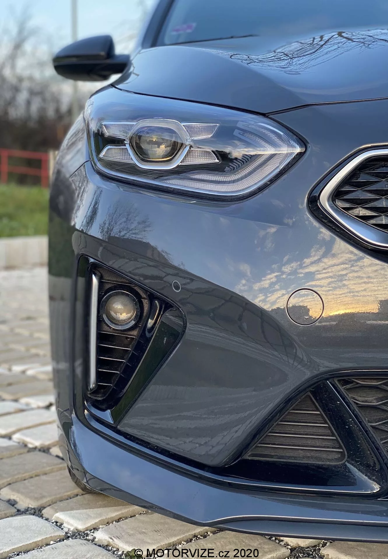 Vue de près et en angle du côté avant gauche d'une Kia Ceed 2018 de couleur sombre, mettant en avant un design de phare élégant avec lumière de jour LED, calandre en nid d'abeille, ensemble de phares antibrouillard et pare-chocs avant aérodynamique, avec un arrière-plan flou et des reflets sur la peinture brillante.