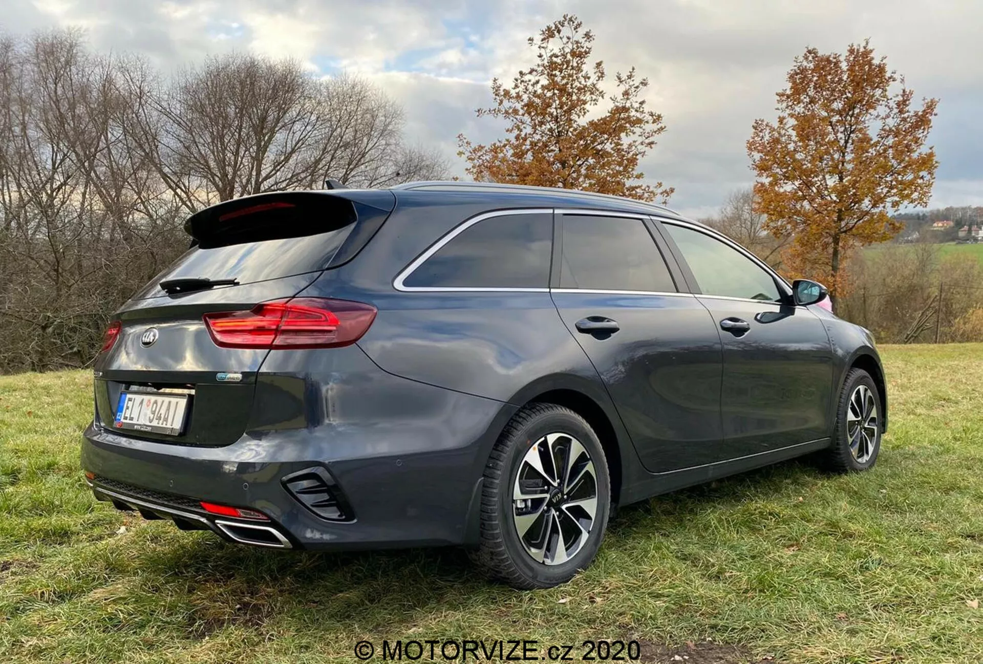Vue arrière trois quarts d'un véhicule moderne de type hatchback ou break, montrant une finition gris foncé, des feux arrière structurés verticalement, un système d'échappement double et des roues en alliage à rayons multiples, garé sur une zone herbeuse avec une légère inclinaison, entouré d'arbres et de cieux nuageux.