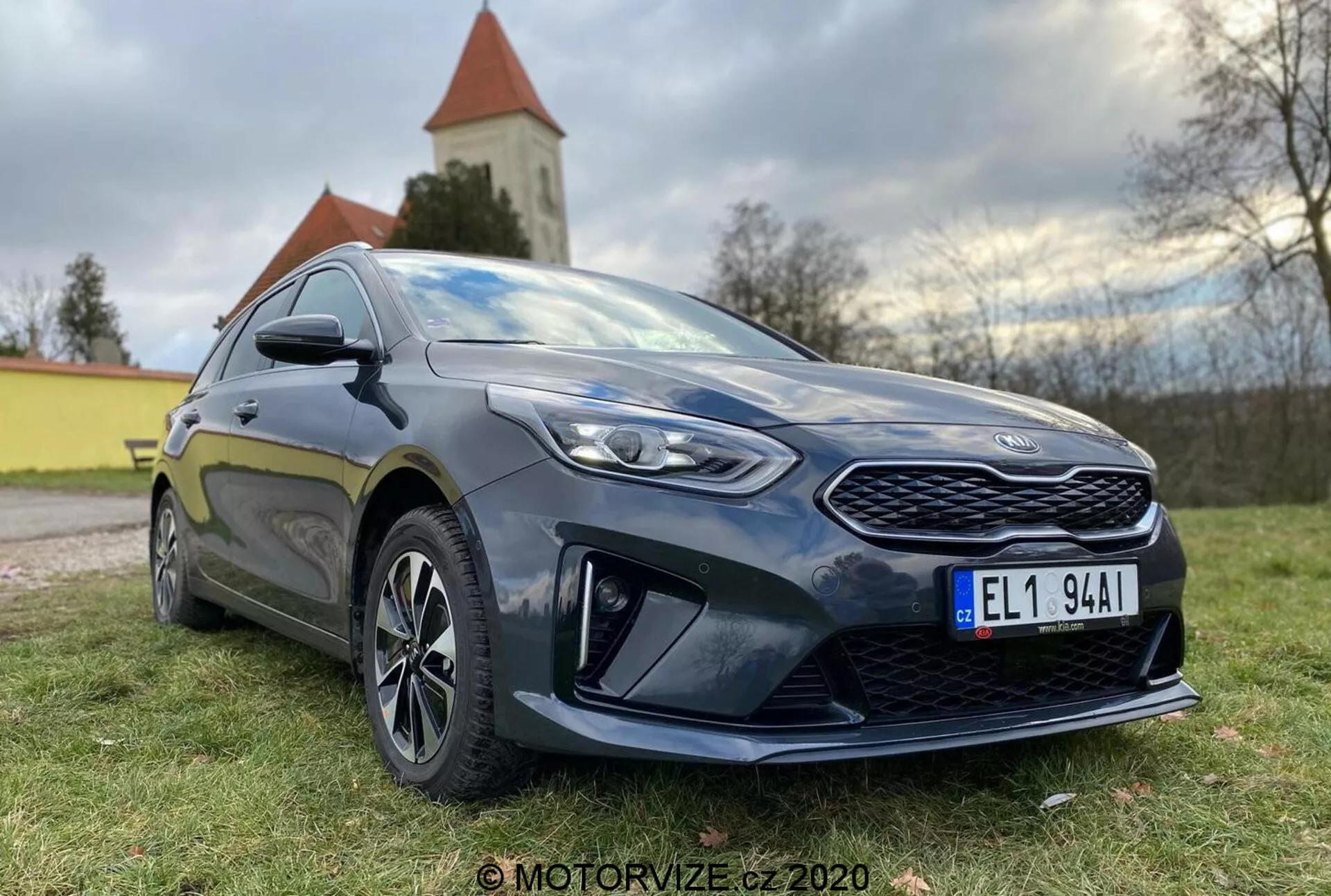 Vue détaillée d'une Kia Ceed 2018 moderne à hayon, photographiée sous un angle avant trois quarts bas, montrant une calandre proéminente, des phares LED angulaires et le profil latéral jusqu'à la porte du conducteur. La voiture est de couleur sombre, probablement bleu foncé ou noir, avec une brillance qui met en évidence son état bien entretenu. Les caractéristiques de design notables incluent une calandre avant à motif, des phares nets avec des lentilles claires, des éléments aérodynamiques prononcés sur le pare-chocs avant inférieur, des jantes alliage à rayons multiples, des rétroviseurs latéraux de couleur carrosserie avec des clignotants intégrés et une jupe latérale sportive. La posture de la voiture suggère un équilibre entre confort et sportivité. La lumière naturelle du jour avec un éclairage doux met en valeur les détails, et un paysage naturel avec une église ou une tour est visible en arrière-plan.