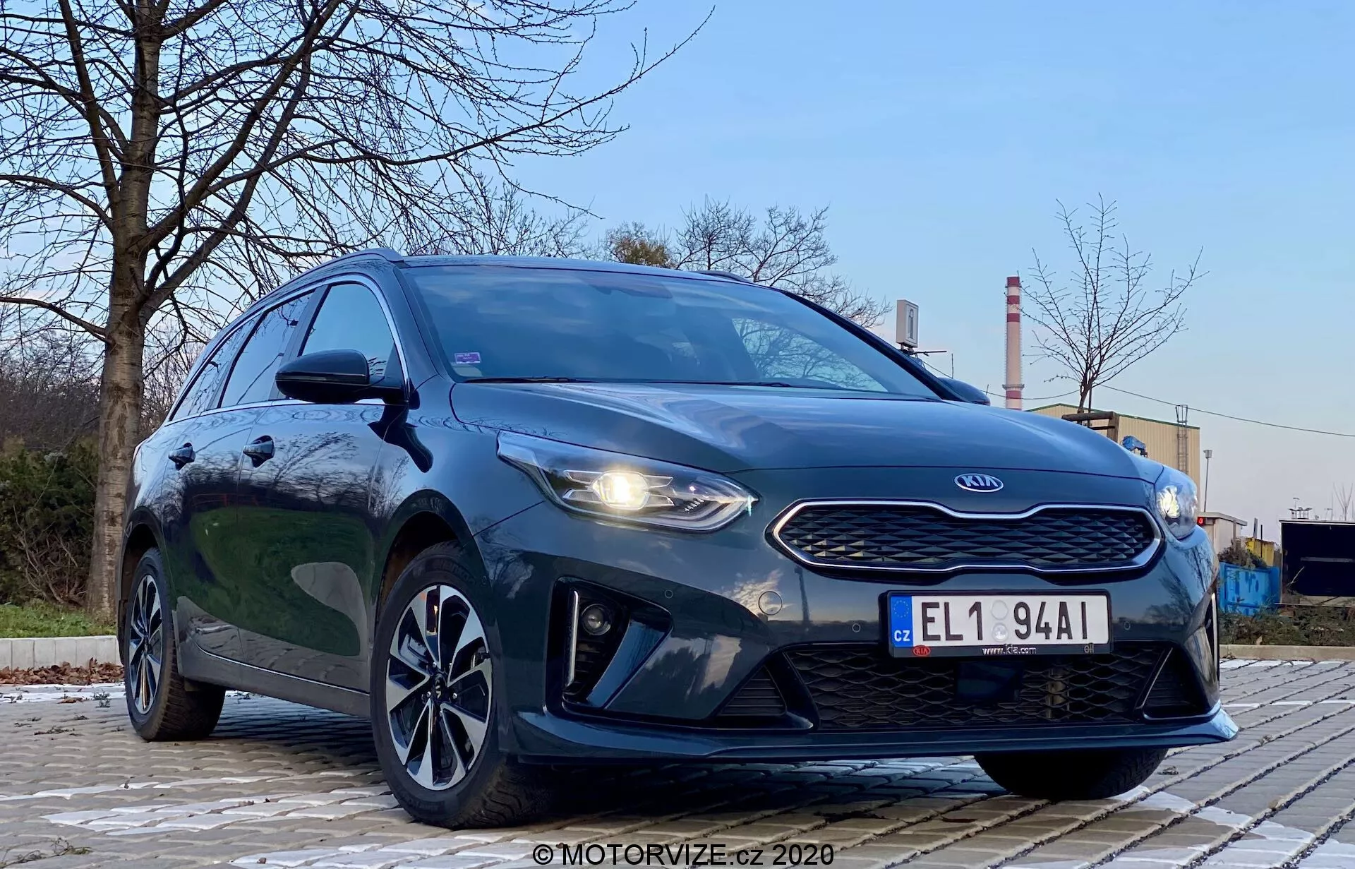 Photographie de la Kia Ceed 2018 prise d'un angle bas avant-gauche, mettant en évidence la calandre, les phares et le design de la face avant du véhicule. Le style agressif de la voiture est accentué par des phares proéminents, une calandre en forme de nez de tigre et des contours de capot. L'image montre également les feux diurnes à LED, le pare-chocs avant aérodynamique et les élégantes jantes en alliage. Le cadre est extérieur avec des arbres et des structures industrielles en arrière-plan, offrant un contraste. La peinture métallique sombre du véhicule est brillante et bien éclairée, avec des reflets indiquant une surface propre. L'image se concentre sur la voiture avec une perspective dramatique à angle bas sous un éclairage extérieur doux.