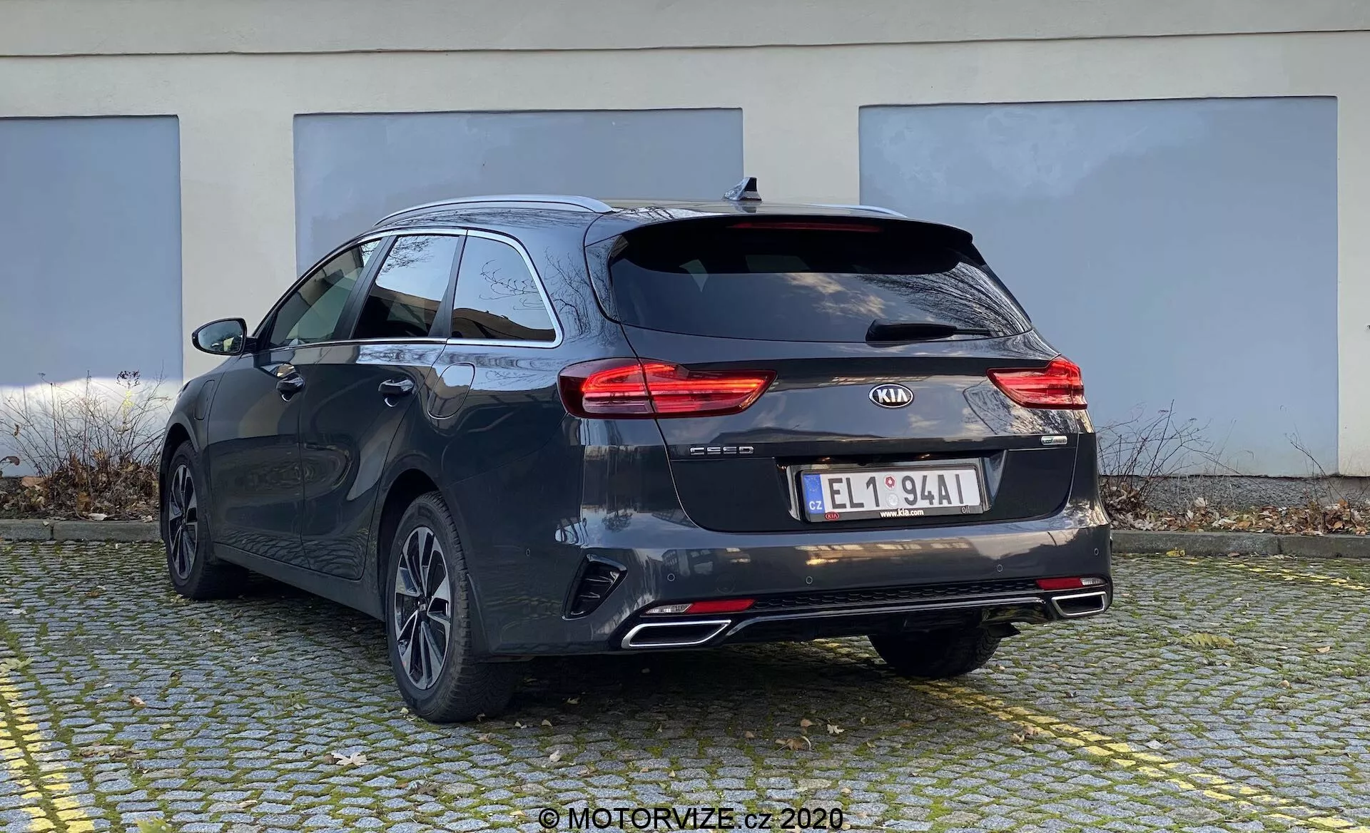 L'image montre une Kia Ceed 2018 en perspective arrière de trois quarts, mettant en évidence l'arrière et le côté du break. La voiture affiche une teinte sombre, peut-être noire ou gris foncé, avec des lignes de carrosserie distinctives et un toit légèrement en pente pour une esthétique sportive. On peut voir les feux arrière à LED modernes, le logo Kia, le nom du modèle 'CEED', des badges supplémentaires, une plaque d'immatriculation de style européen, des roues arrière en alliage multibranches, des doubles sorties d'échappement et un essuie-glace arrière. Il est probable qu'il y ait une caméra de recul et des capteurs de stationnement non visibles. Le petit becquet de toit et les barres de toit indiquent des options de chargement supplémentaires. La voiture est stationnée sur une zone pavée avec un bâtiment en arrière-plan, montrant le profil arrière et partiellement celui du côté depuis un angle légèrement bas.