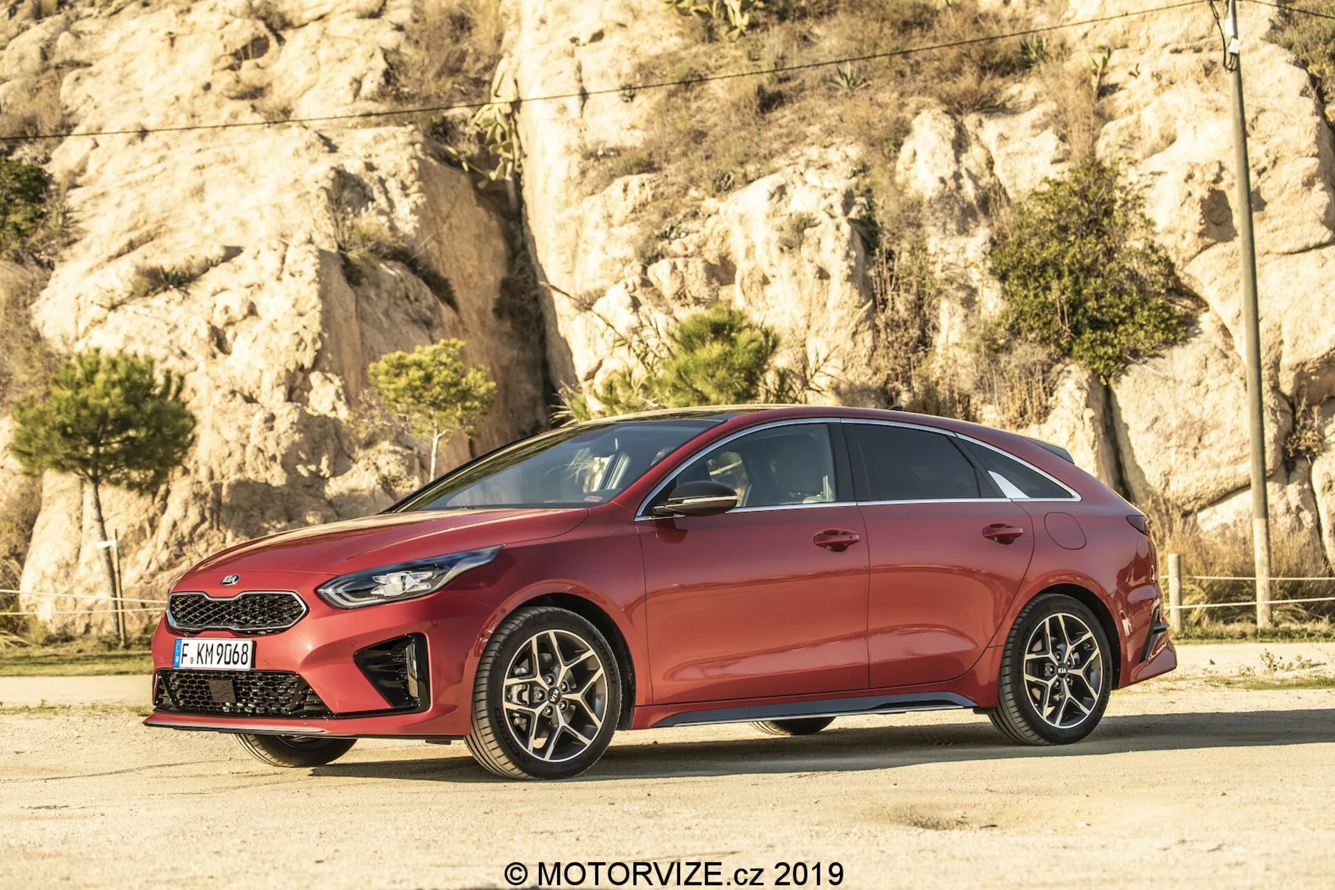 Vue de profil latéral d'une Kia Ceed 2021 rouge, avec un design élégant et aérodynamique, des lignes de toit inclinées et des lignes profilées sur les portes. La voiture, un modèle à cinq portes, possède des garnitures de fenêtre chromées et des montants B noirs créant un effet de panneau de verre continu. Les jantes en alliage à plusieurs rayons, avec des freins à disque visibles, ajoutent une touche sportive. Les phares acérés et la grille partiellement visible marquent l'avant, tandis que les rétroviseurs de couleur carrosserie avec clignotants intégrés améliorent la visibilité et la sécurité.