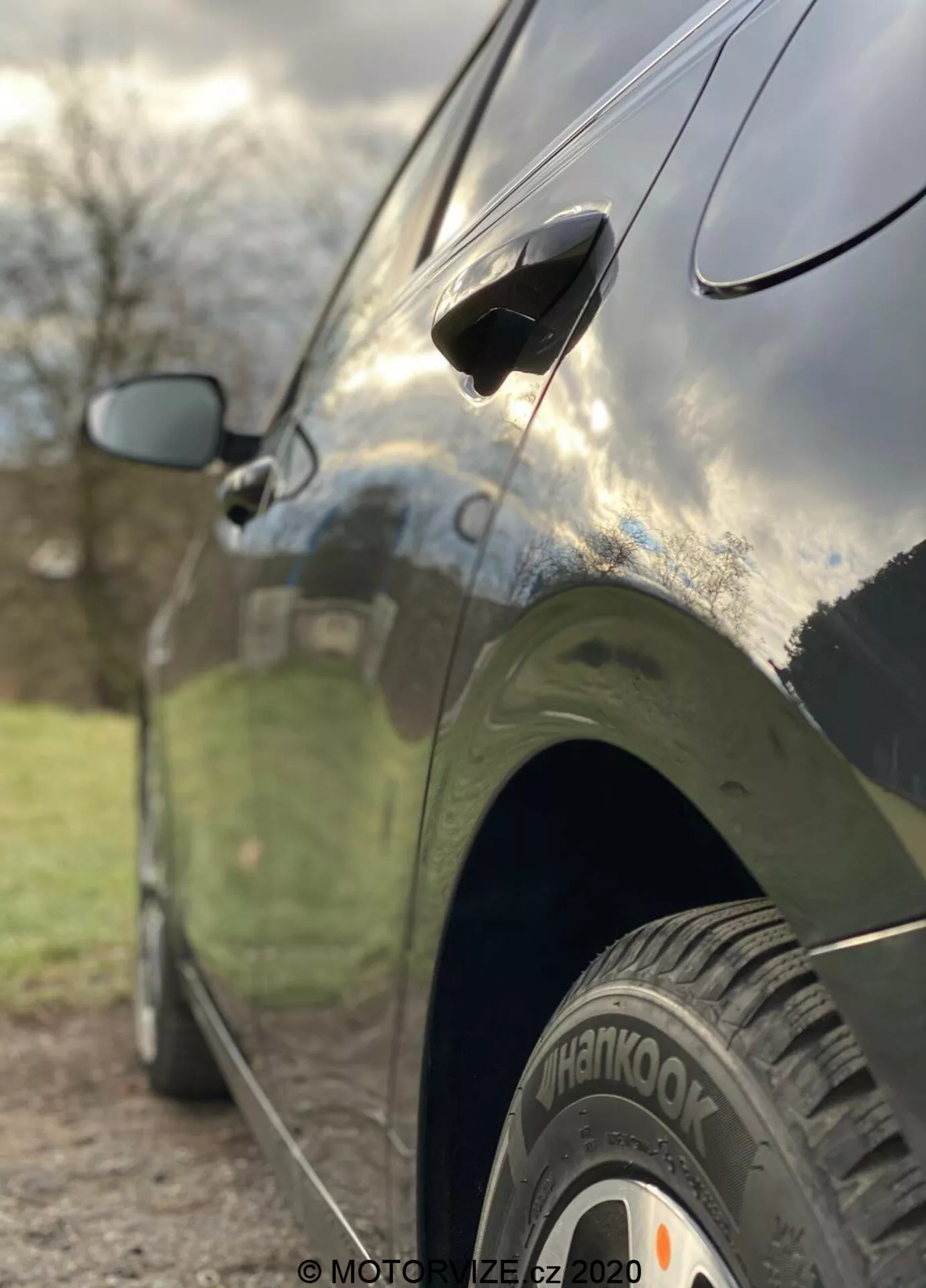 Vue de côté en gros plan de la Kia Ceed 2018, mettant en valeur des caractéristiques telles que la roue arrière avec le pneu 'Hankook', la poignée de porte aérodynamique et le rétroviseur latéral partiellement flou. La photo est prise d'un angle bas avec une faible profondeur de champ qui met en nette focale la roue arrière et le bas de la porte de la voiture, tandis que l'arrière-plan est artistiquement flou. La voiture a une finition sombre et brillante qui reflète le ciel et les environs, montrant des lignes de design élégantes et une carrosserie bien entretenue.