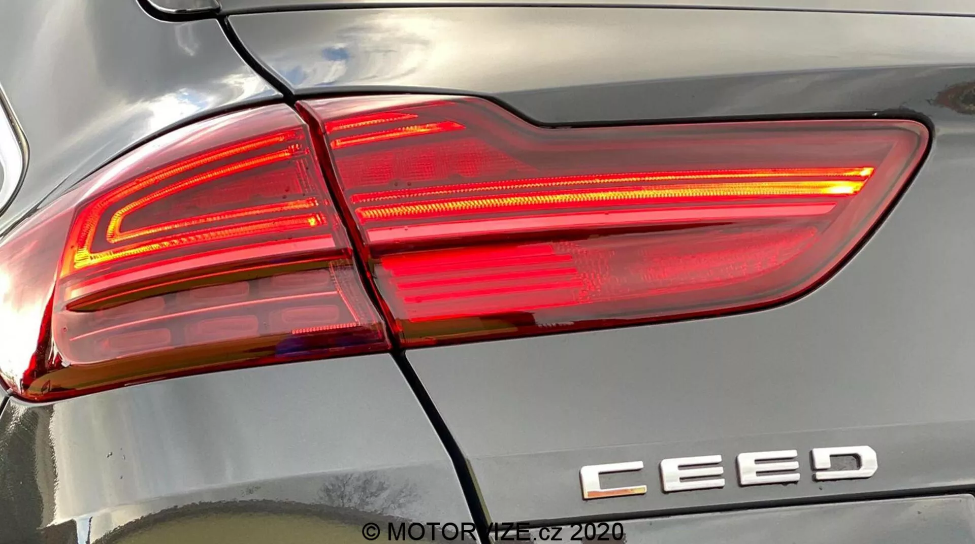 Vue détaillée de près de l'ensemble du feu arrière droit d'une Kia Ceed 2018, illuminé, montrant un design moderne avec des lignes nettes et angulaires, des sections de lentilles claires et rouges, et le badge du modèle 'CEED'. Une ligne horizontale sépare le feu arrière du pare-chocs arrière. Les reflets sur la carrosserie soulignent la courbure de la surface. Filigrane 'MOTOR1.cz ©2020'.