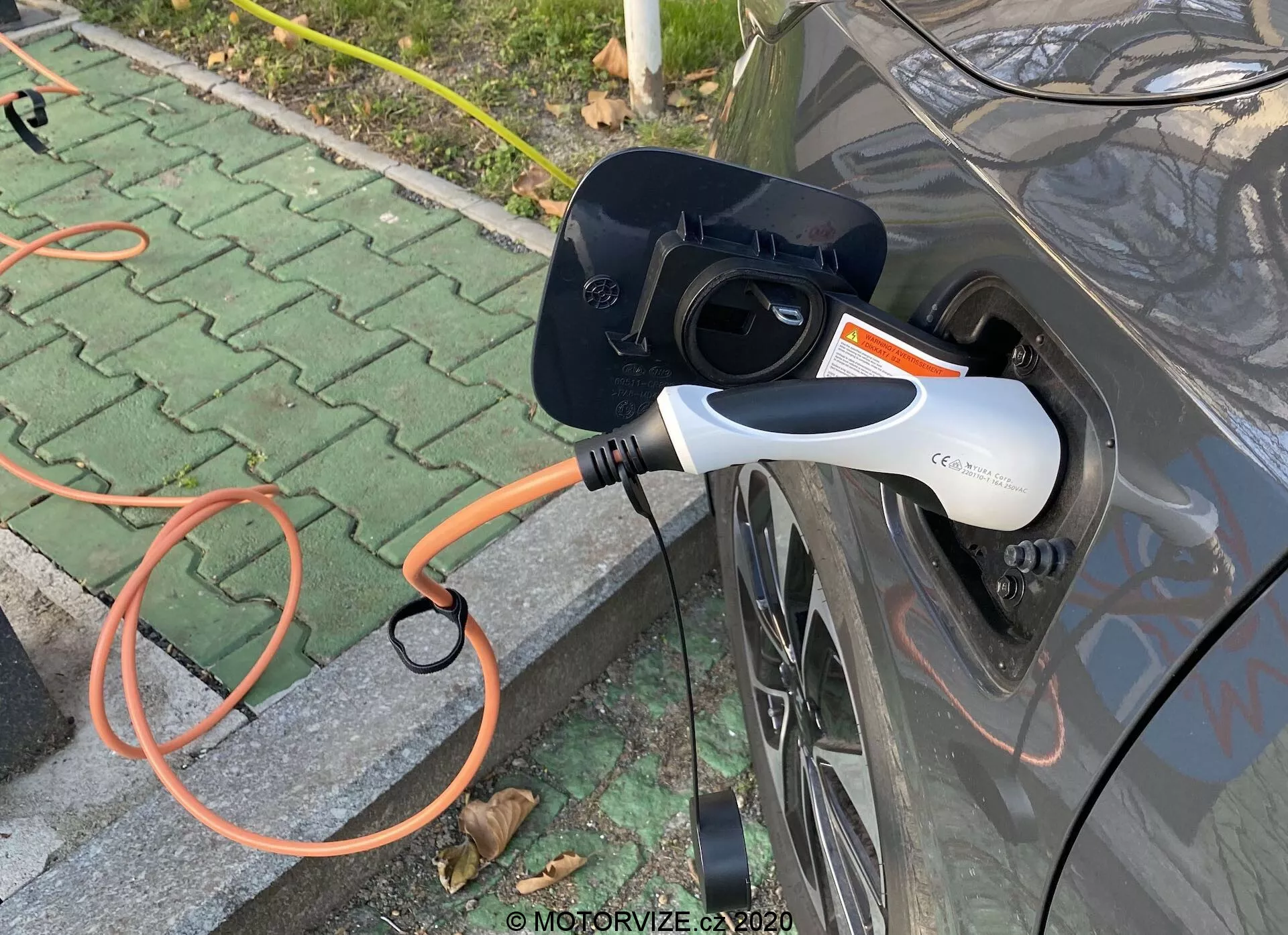 Vue latérale rapprochée d'un véhicule électrique (EV) gris en cours de charge, mettant en évidence le port de charge ouvert et un connecteur EVSE orange branché à la voiture. Le connecteur de charge moderne blanc et noir est équipé d'une lumière indicatrice LED. L'image se concentre sur l'interaction de chargement, située dans une zone pavée extérieure avec quelques feuilles éparses.