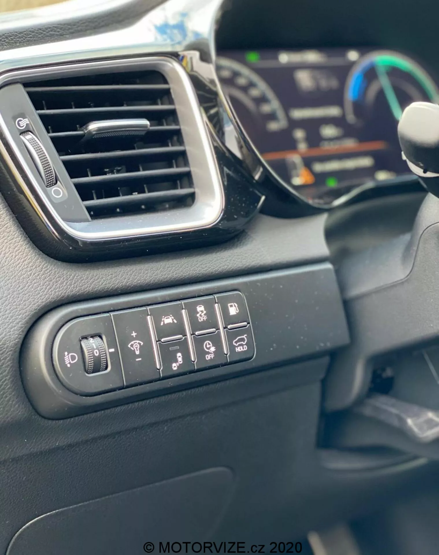 Vue rapprochée de l'intérieur de la Kia Ceed 2018 montrant les boutons de commande pour le réglage des rétroviseurs, le chauffage de la lunette arrière, le chauffage du volant, l'avertissement de franchissement de ligne, l'assistance au stationnement et le contrôle de traction. L'image présente également une élégante bouche d'aération, une partie du tableau de bord numérique en arrière-plan flou et le bord du volant. Les matériaux intérieurs sont principalement des plastiques durs avec une finition mate, capturés à la lumière du jour.