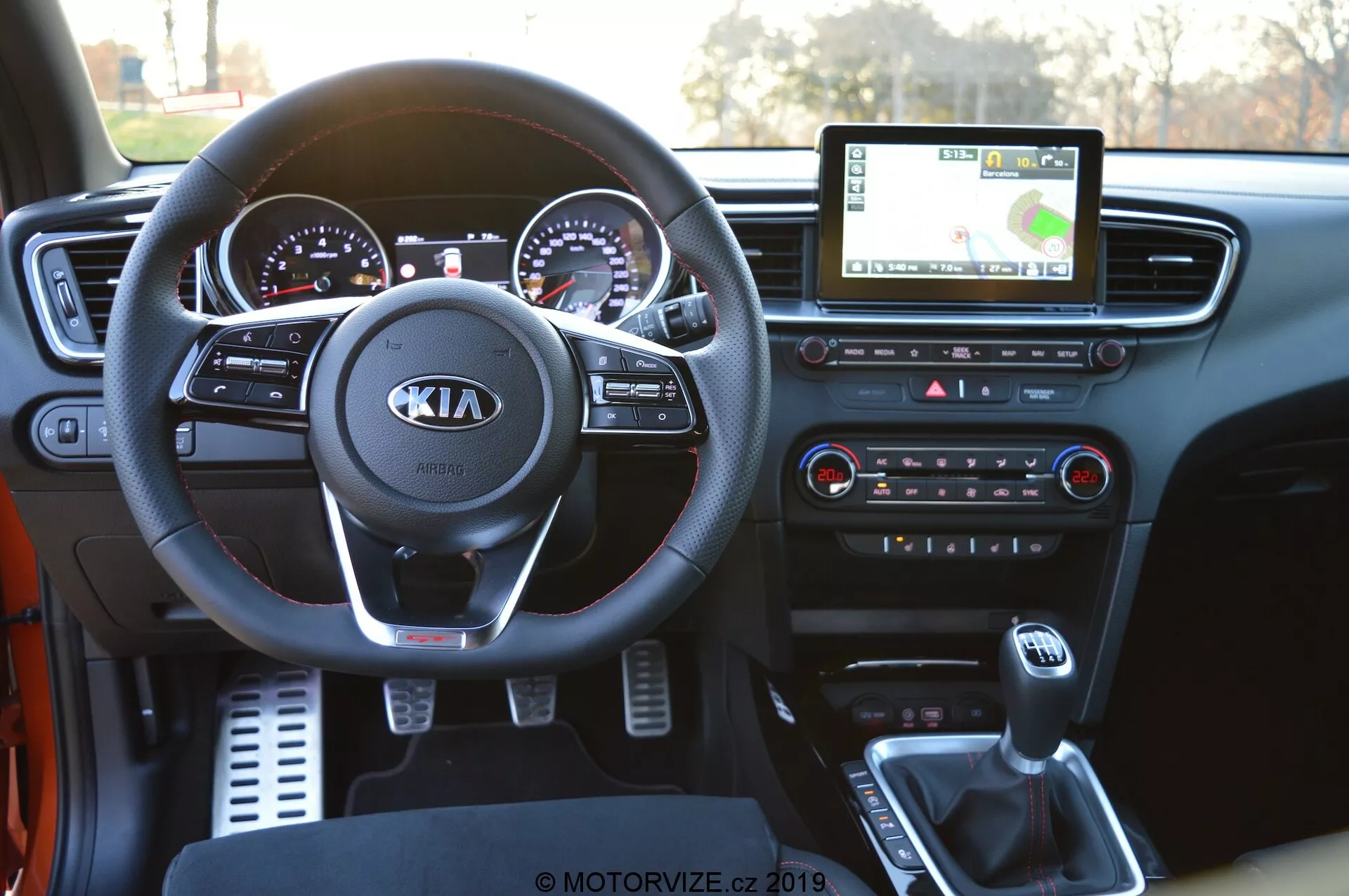 Vue intérieure d'une Kia Ceed 2021 depuis la perspective du conducteur, présentant le tableau de bord détaillé. L'image met en évidence le volant avec le logo Kia et les commandes intégrées pour l'audio, les appels et le régulateur de vitesse. Derrière, le tableau de bord avec des jauges analogiques accentuées de chrome et un écran numérique est visible. L'écran tactile central de l'info-divertissement affiche la navigation GPS, tandis qu'en dessous se trouvent les boutons et les boutons de commande du climat. La console centrale montre le levier de changement de vitesse, les porte-gobelets et un port USB pour la connectivité des appareils, mettant en avant la technologie moderne embarquée dans le véhicule.