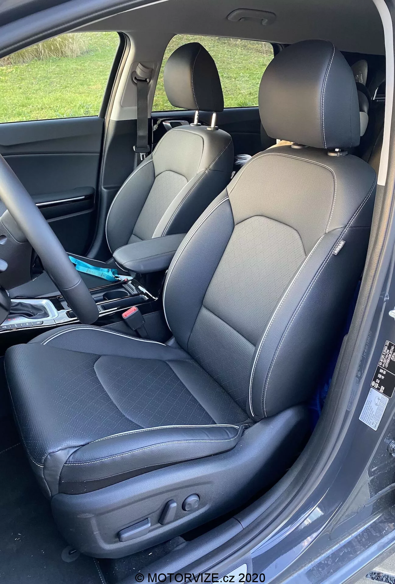 Gros plan de l'intérieur de la Kia Ceed 2018 montrant les sièges conducteur et passager avec une sellerie en cuir bicolore et des coutures contrastantes, un design de siège baquet avec des renforts latéraux, et des réglages électriques sur le siège conducteur pour le contrôle de l'inclinaison et de la position. Des plis légers indiquent une utilisation et divers objets sont visibles sur le siège passager.