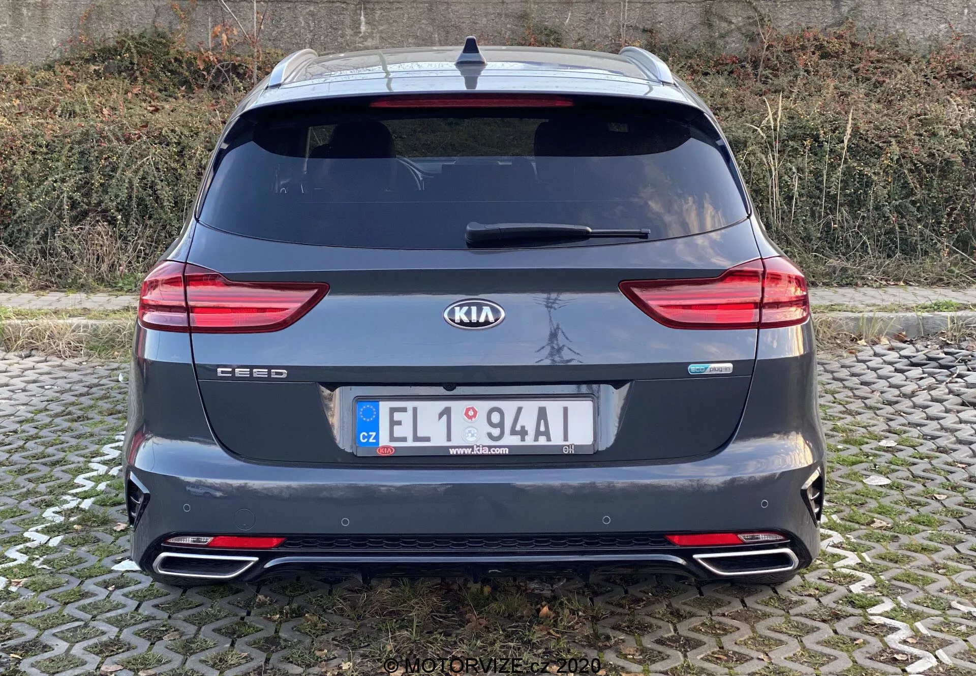 Vue arrière de la Kia Ceed 2018 montrant toute la section arrière avec les badges de la marque et du modèle, des feux arrière LED modernes, la lunette arrière, des embouts d'échappement doubles, le pare-chocs arrière avec des réflecteurs, un spoiler de toit, une antenne et des capteurs de stationnement arrière. L'image est claire, prise à hauteur des yeux avec un éclairage naturel dans des conditions nuageuses.