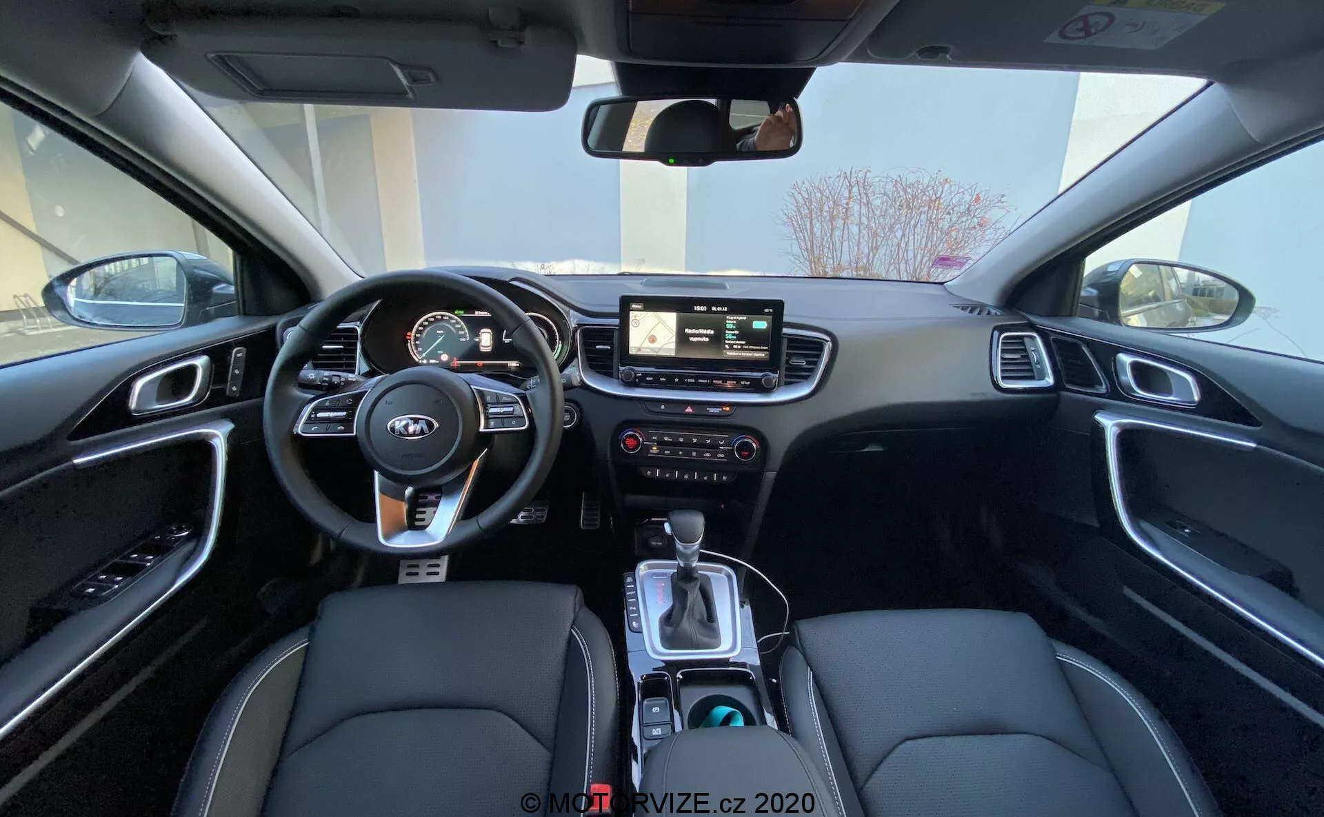 Vue grand angle de l'intérieur de la Kia Ceed 2018 depuis la banquette arrière, montrant la zone du conducteur, y compris le volant avec commandes intégrées, le tableau de bord avec instruments numériques et analogiques, le système d'infodivertissement central avec écran tactile, les commandes manuelles pour l'infodivertissement et le climat, et la console centrale avec levier de vitesse, sélecteur de mode de conduite, porte-gobelets et un intérieur noir avec des sièges en cuir synthétique avec surpiqûres blanches et des garnitures en aluminium brossé.