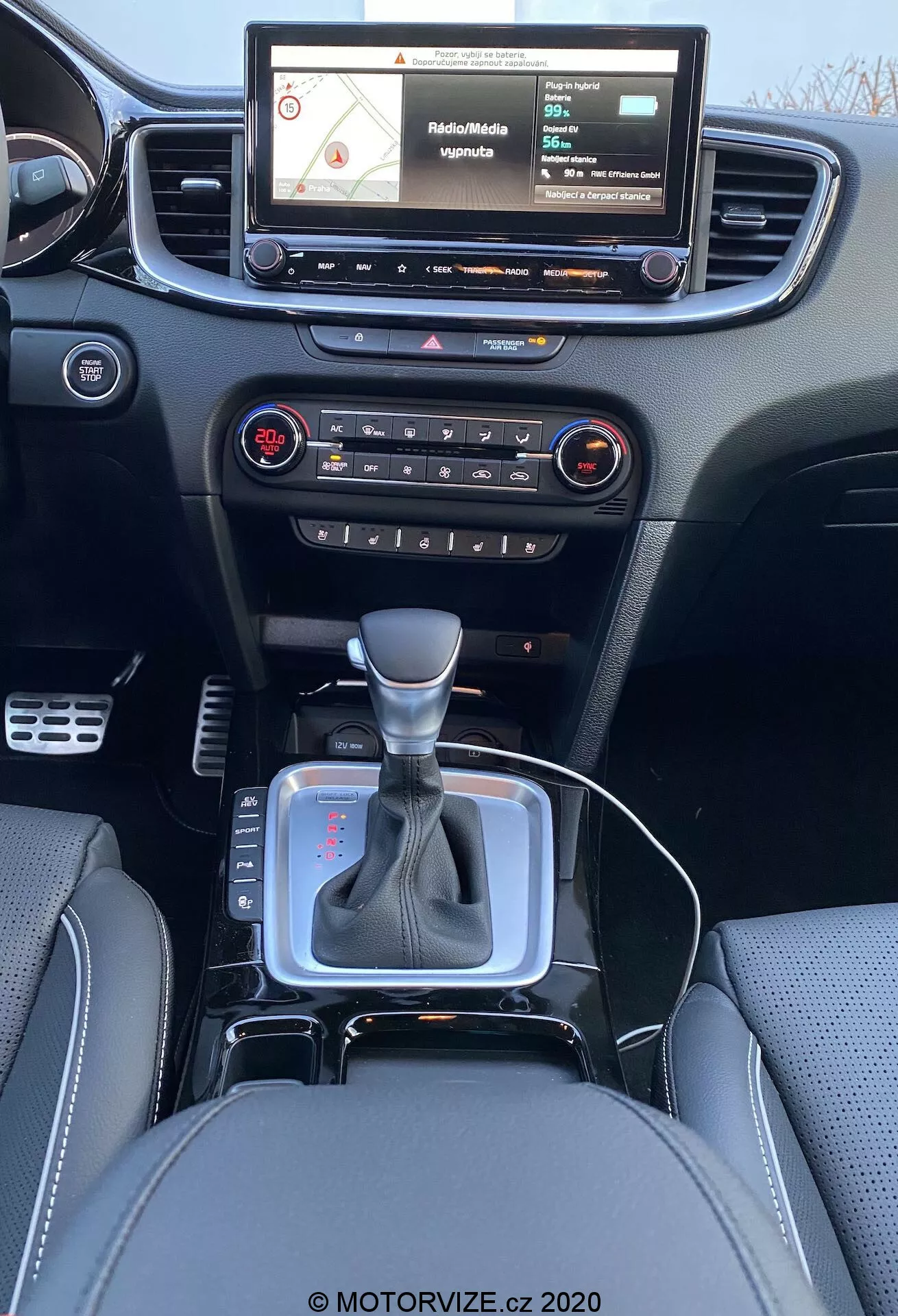 Vue intérieure d'une Kia Ceed 2018 depuis le siège arrière, affichant un tableau de bord moderne avec un système d'infodivertissement à écran tactile montrant une carte de navigation, des boutons physiques et des boutons pour le contrôle audio et climatisation, une bouche d'aération centrale, un levier de vitesse en position neutre avec un accent argenté et une gaine en cuir noir, un petit espace de rangement près du levier de vitesse probablement pour une prise 12V ou des ports USB, des sièges en cuir avec des coutures détaillées.