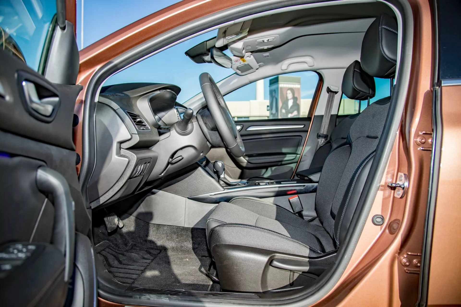 Vue intérieure de la Kia Ceed Pro-Cee'd 2009 à travers la porte du conducteur ouverte montrant les sièges avant en tissu foncé, commandes intégrées au volant, tableau de bord partiellement visible avec écran d'infodivertissement, console centrale avec levier de vitesse, tapis de sol voiture et schéma de couleur bicolore