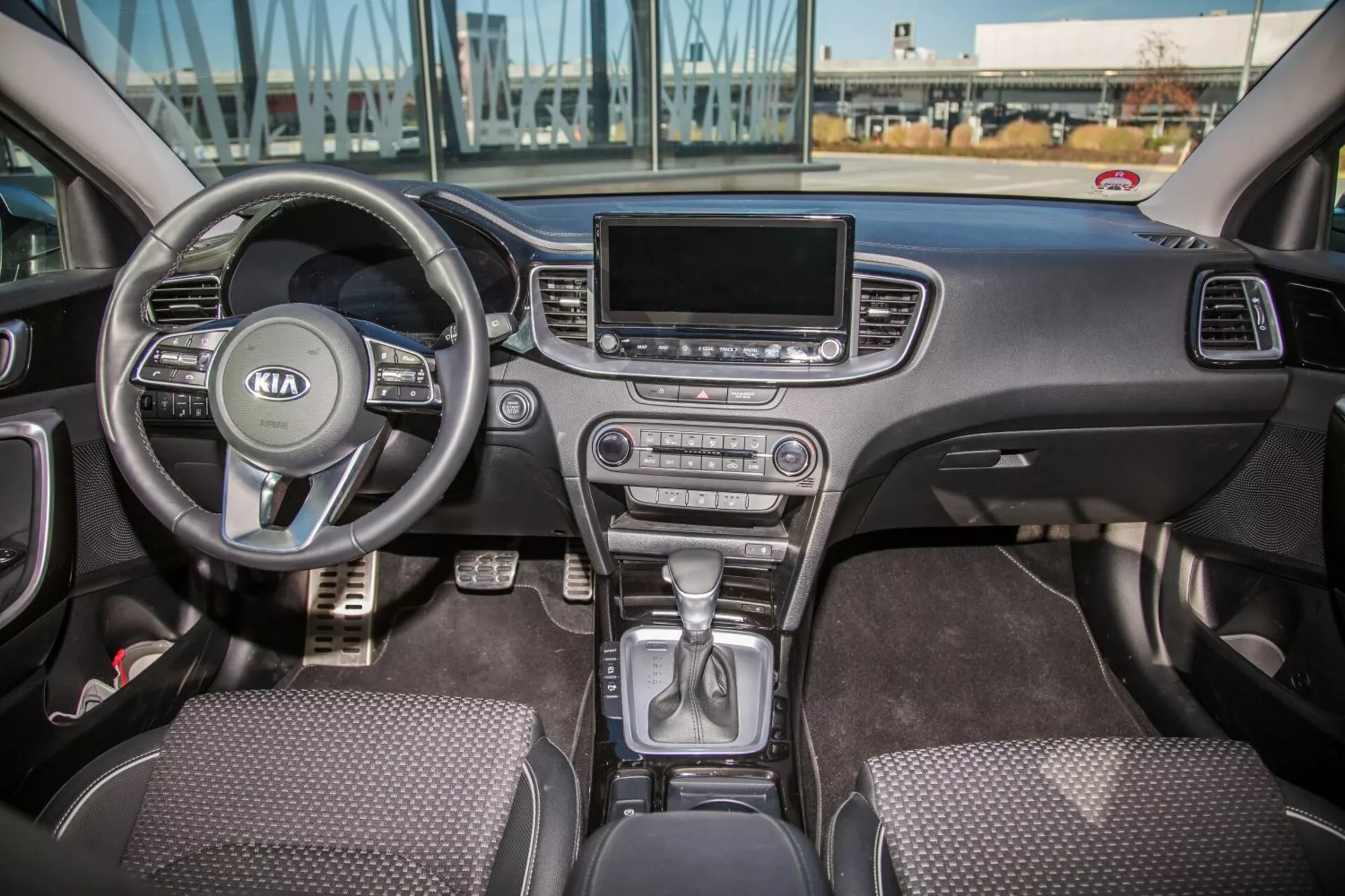 Vue intérieure de la Kia Cee'd 2009 montrant la cabine avant complète incluant le volant multifonctions avec logo Kia, cluster d'instruments derrière, levier de vitesse en position de parking sur la console centrale, boutons de commande climatique, écran d'infotainment, sièges avec revêtement en tissu, et contrôles de portes conducteur et passager à la lumière naturelle du jour