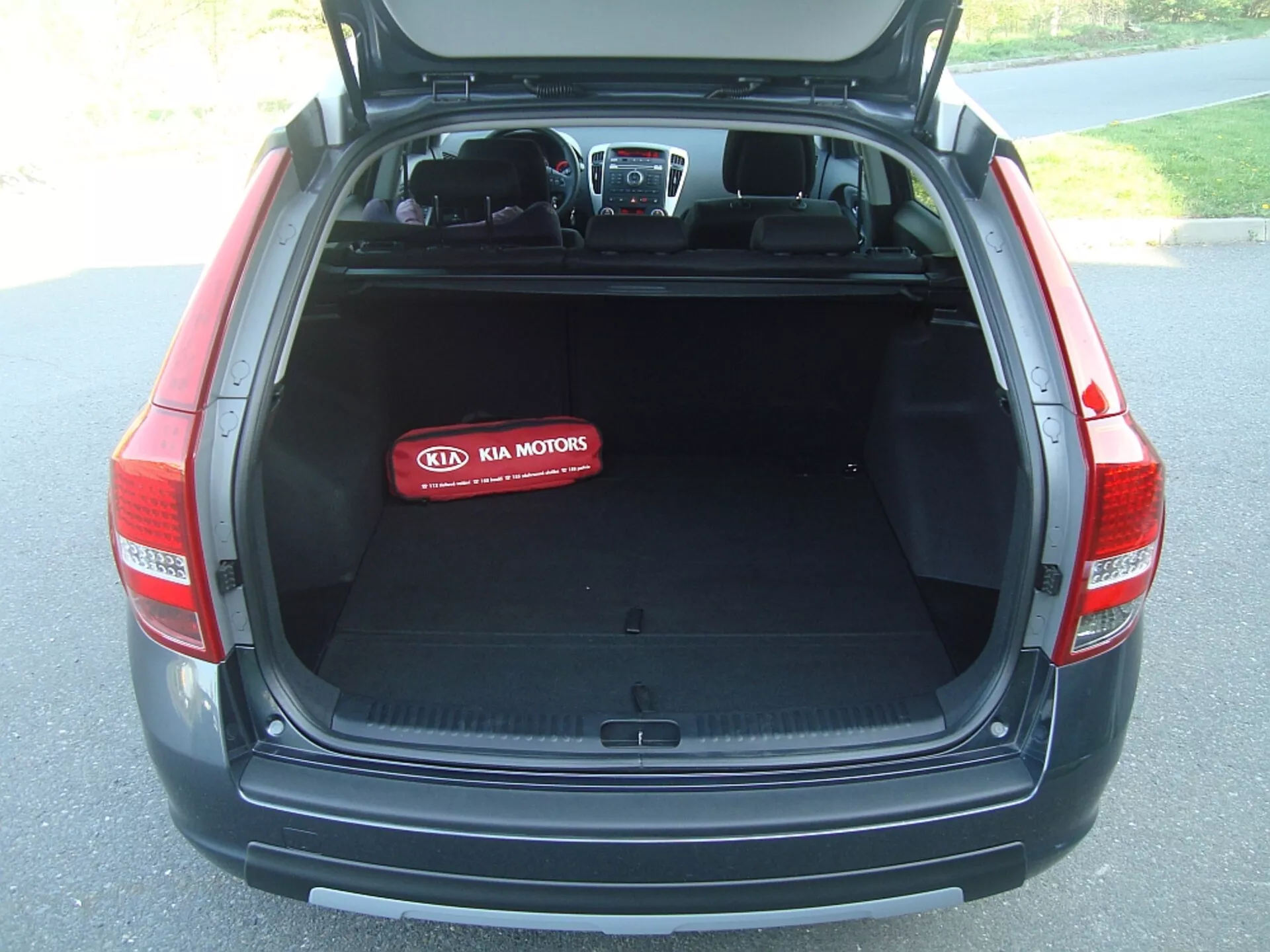 Section arrière du coffre du hatchback Kia Cee'd de 2009 avec le couvercle ouvert révélant un espace de chargement spacieux et vide, couverture rétractable, tapisserie intérieure en textile et logo Kia Motors sur une couverture protectrice rouge, mettant en évidence les caractéristiques de conception, de sécurité et de technologie du véhicule.