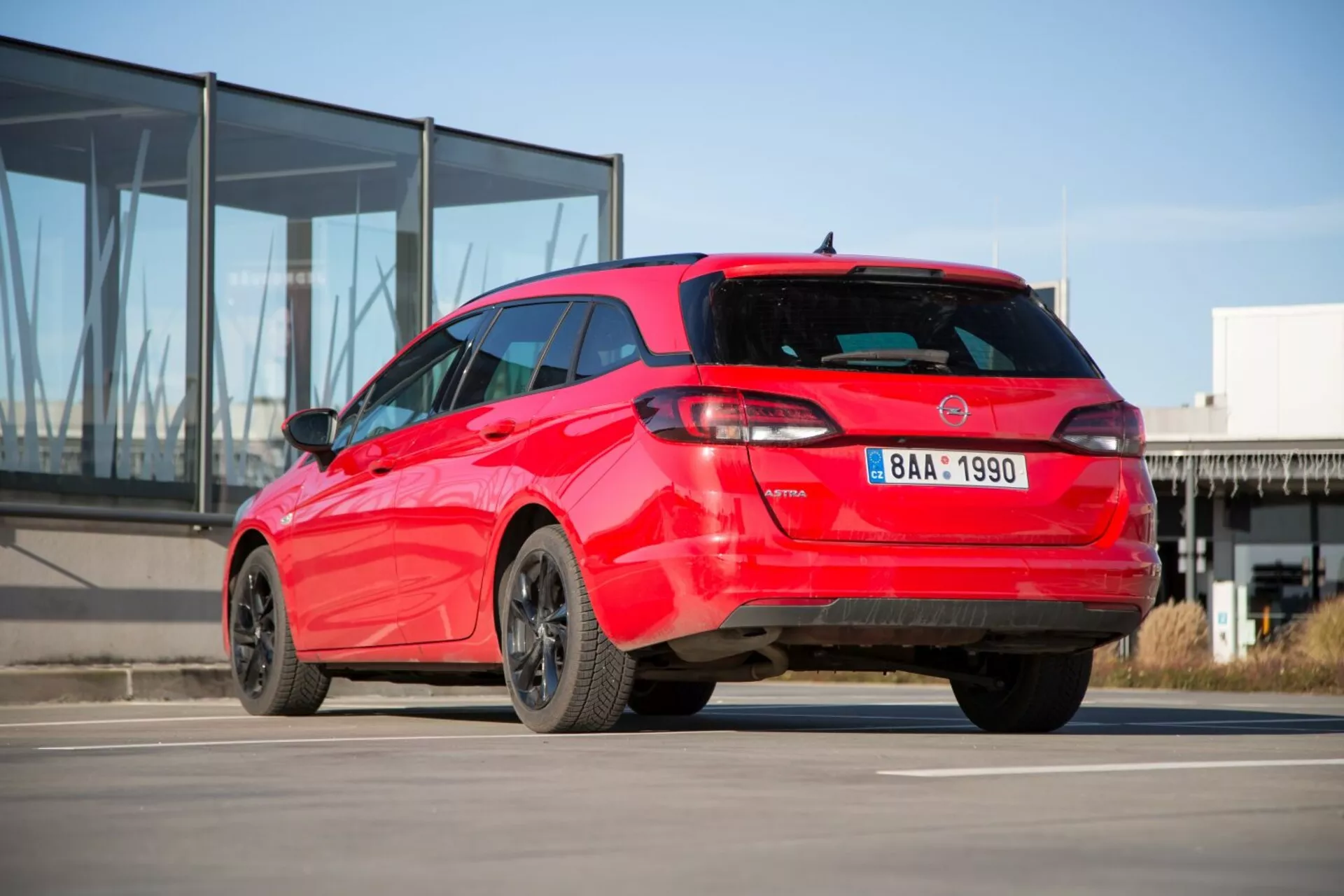 Kia Ceed hatchback 2009 en rouge présentant sa vue arrière trois quarts sportive sur béton avec un bâtiment moderne à façade en verre en arrière-plan, montrant un design distinctif des feux arrière, un spoiler de toit, antenne aileron de requin, jantes en alliage et éventuels capteurs de stationnement.
