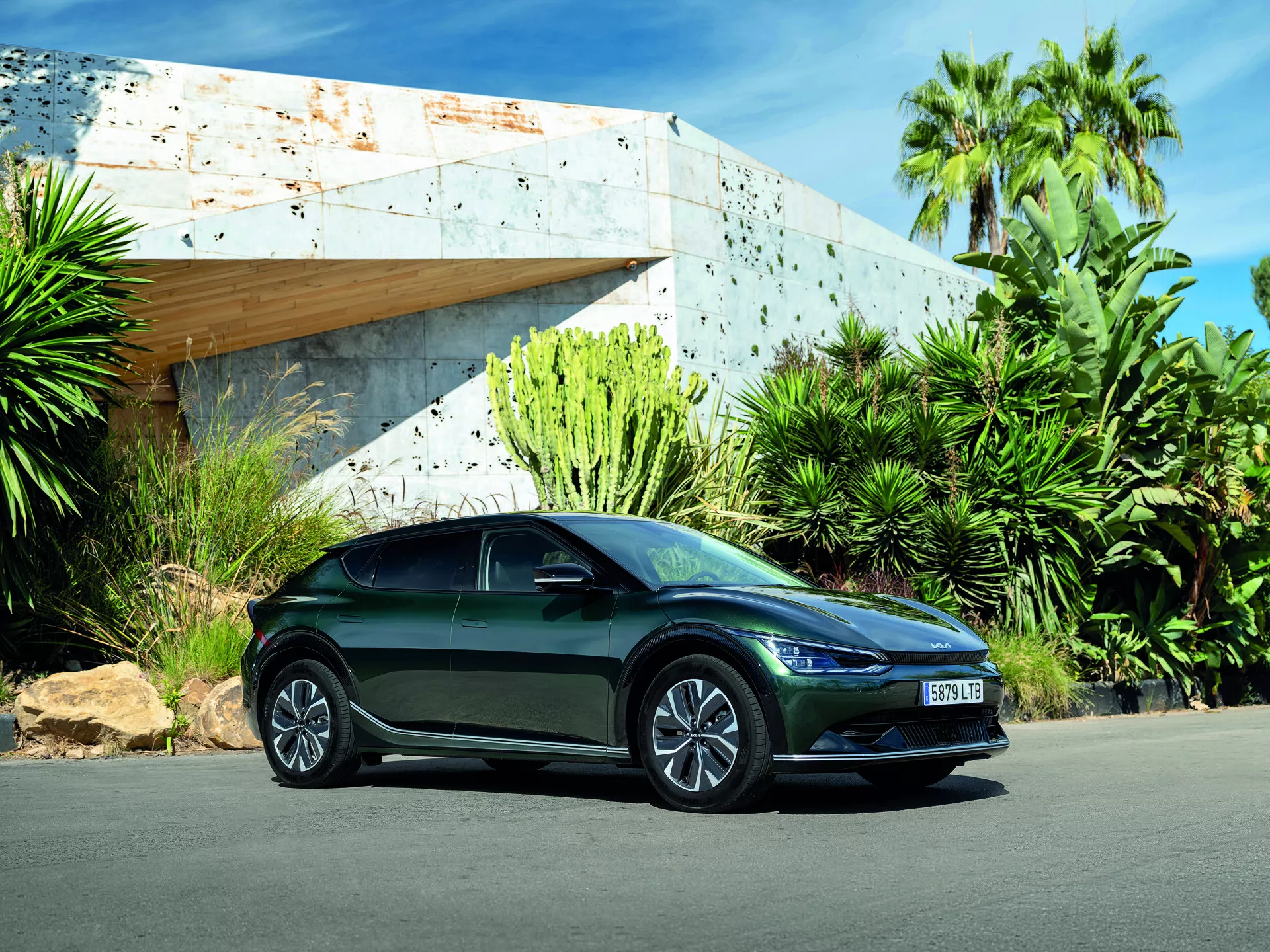 Kia EV6 2021 capturée sous un angle bas soulignant son design élégant, sa posture sportive avec une vue de trois quarts avant-côté montrant les phares LED, le corps aérodynamique, les roues en alliage, les clignotants intégrés sur les rétroviseurs latéraux, garée en extérieur sous une lumière du jour vive avec un bâtiment moderne et une verdure luxuriante en arrière-plan